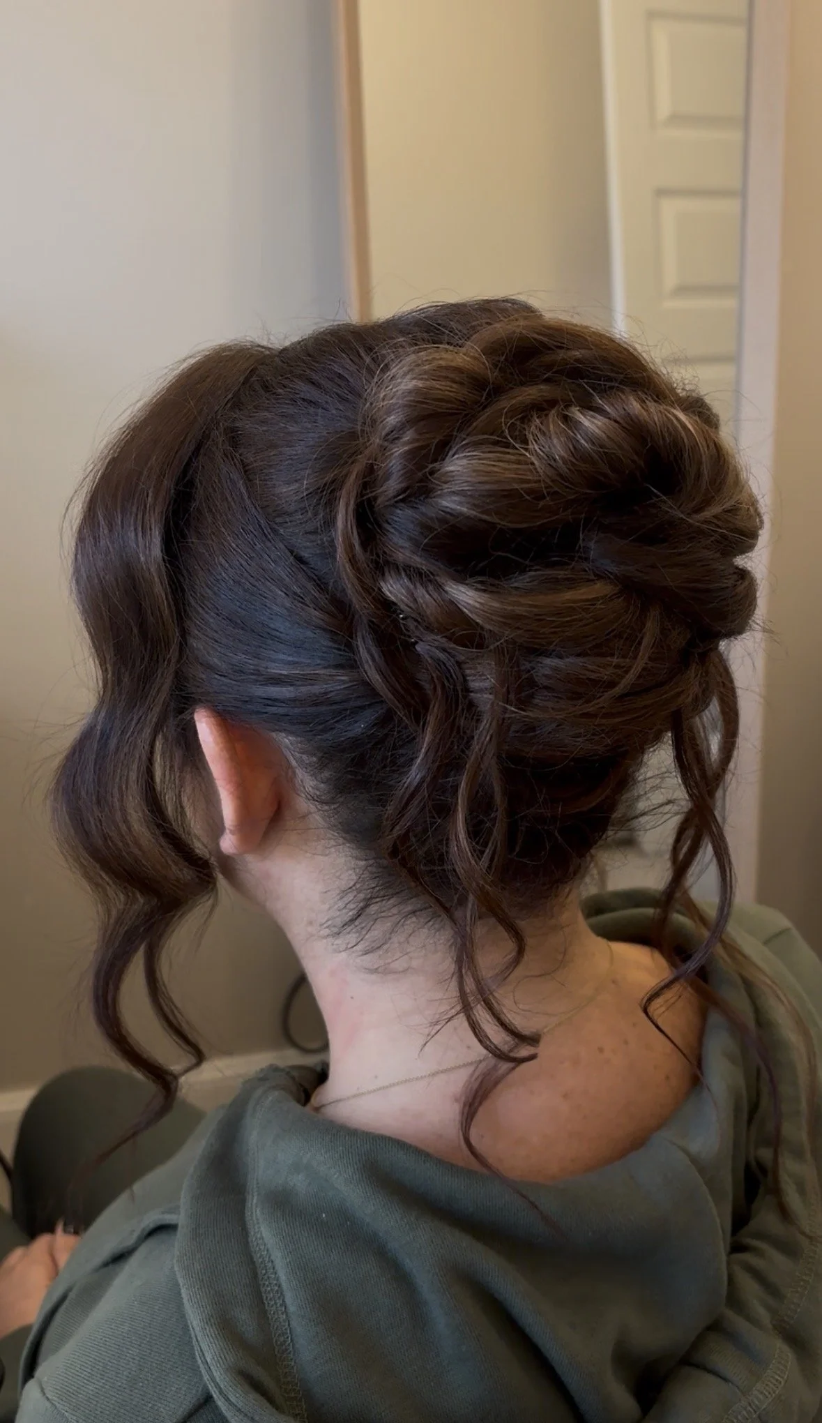 A woman with dark, curly hair styled in an elegant updo with loose curls hanging down the back and sides after a hairstyle appointment.