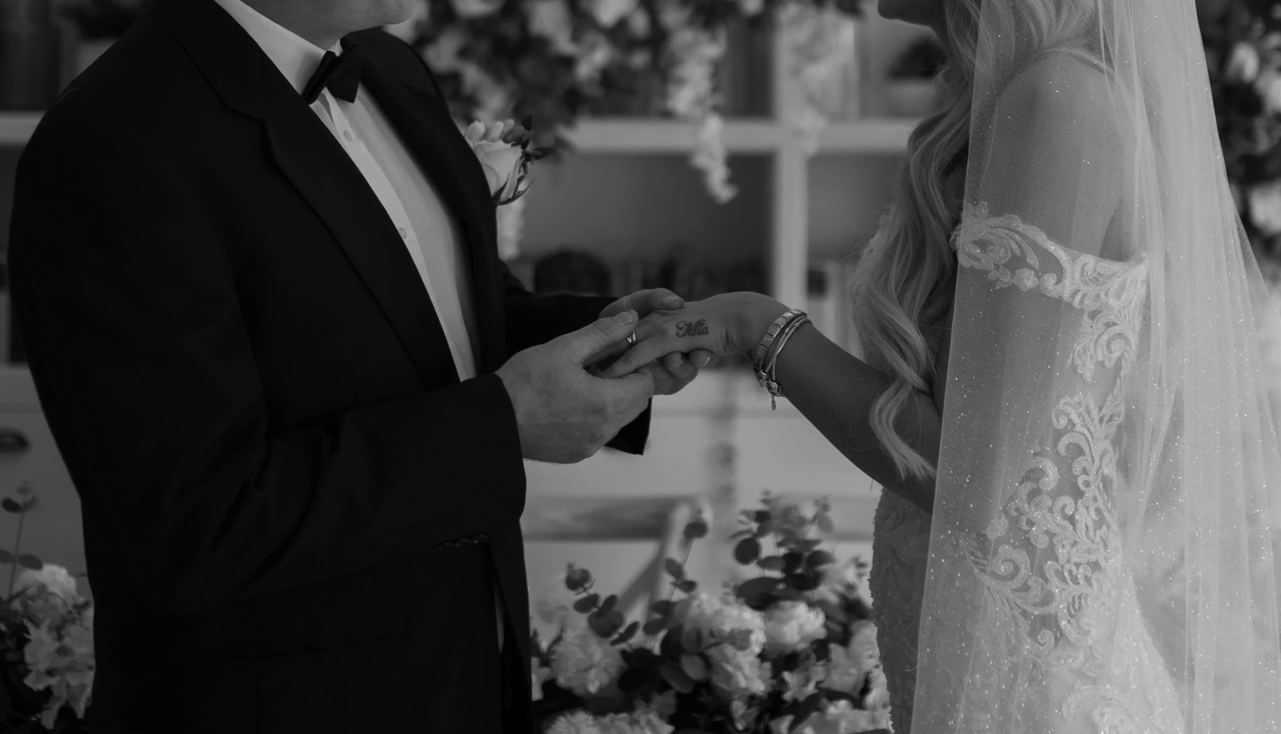 A bride and groom holding hands during a wedding ceremony, with the groom wearing a tuxedo and the bride in a detailed wedding gown and veil.