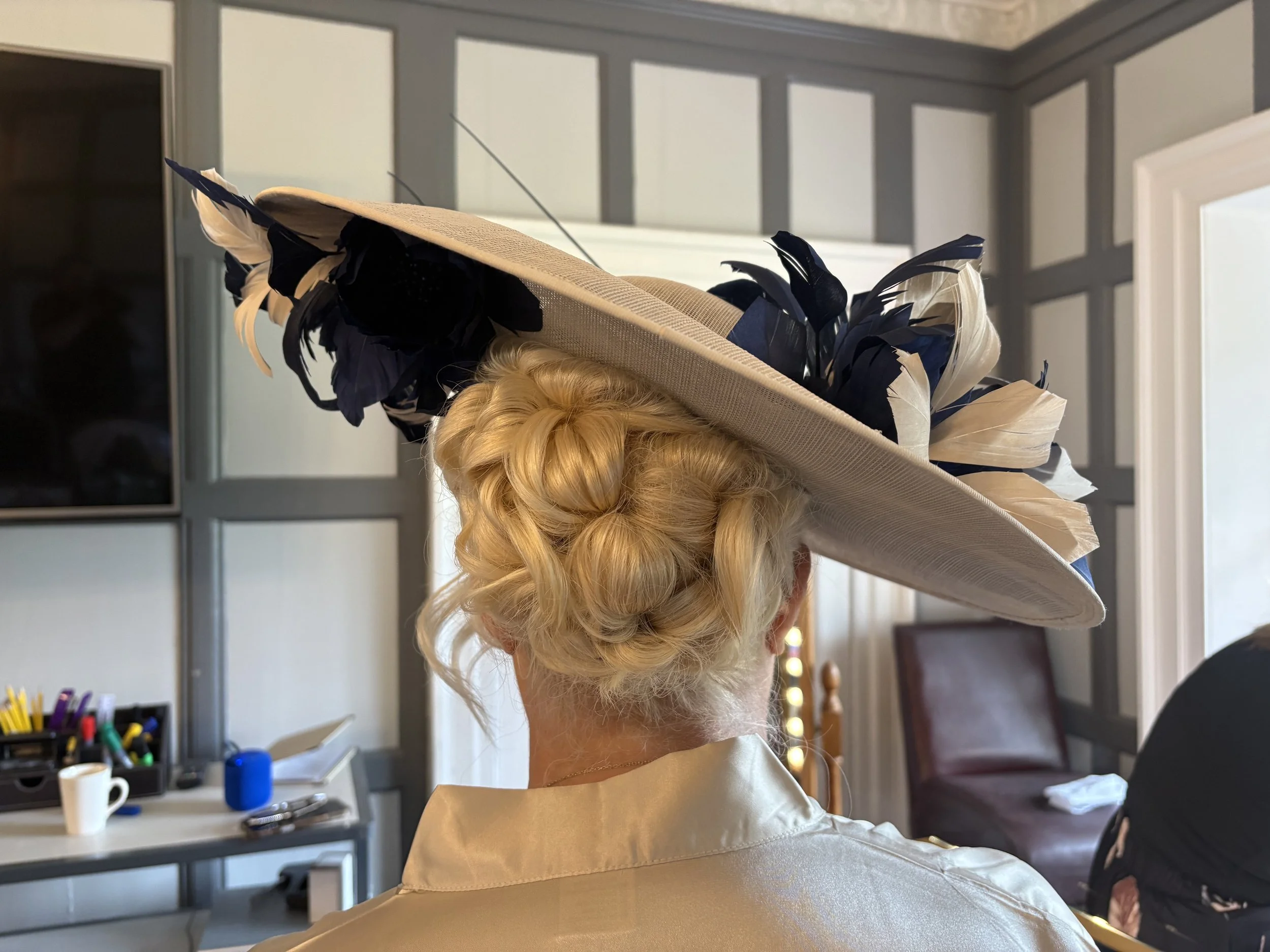 A person with curly blonde hair in a bun wearing a wide-brimmed beige hat with black and beige feathers, in an indoor setting with a table and office supplies in the background.