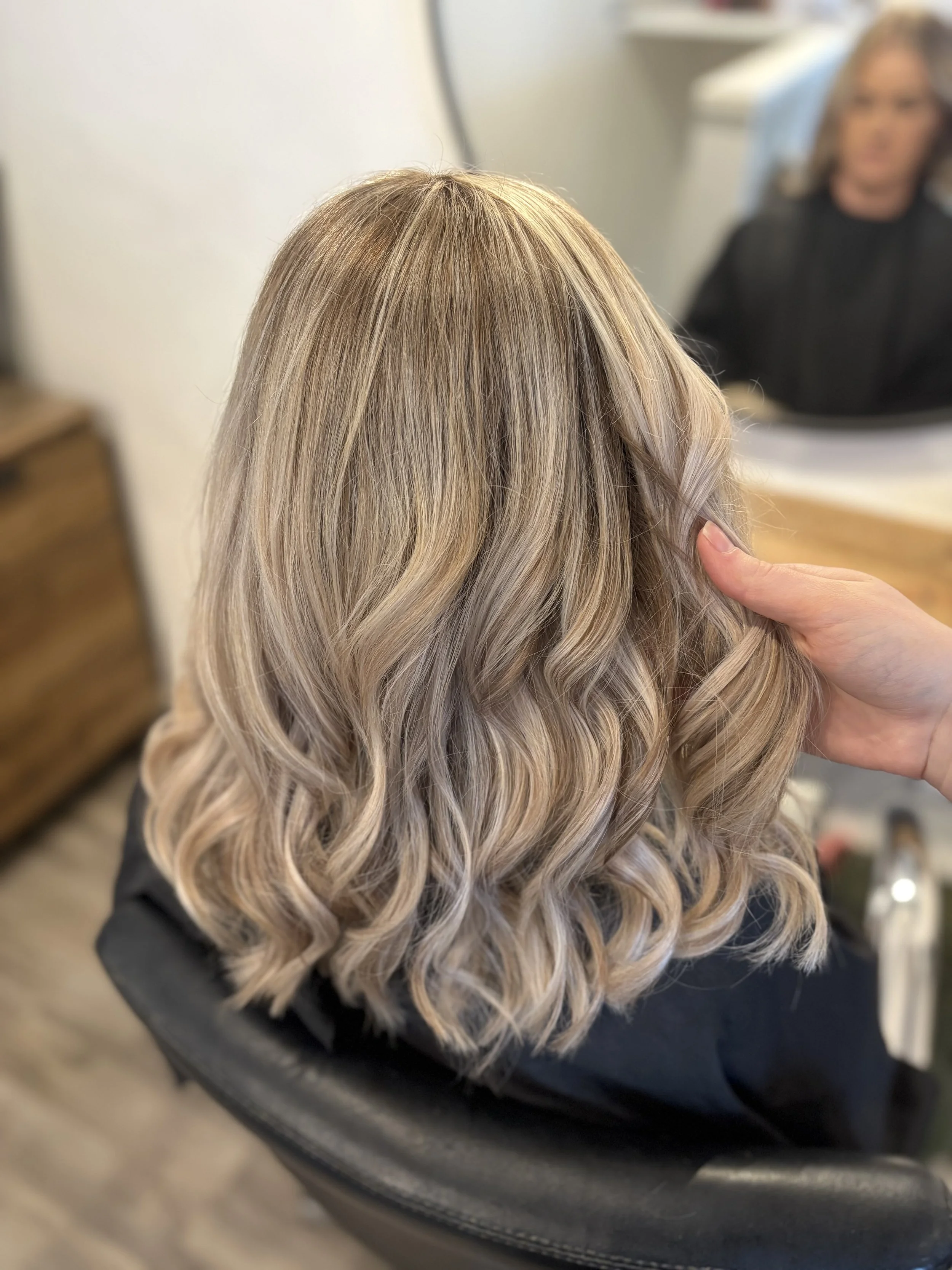 Close-up of a woman with long, blonde, wavy hair in a salon mirror.