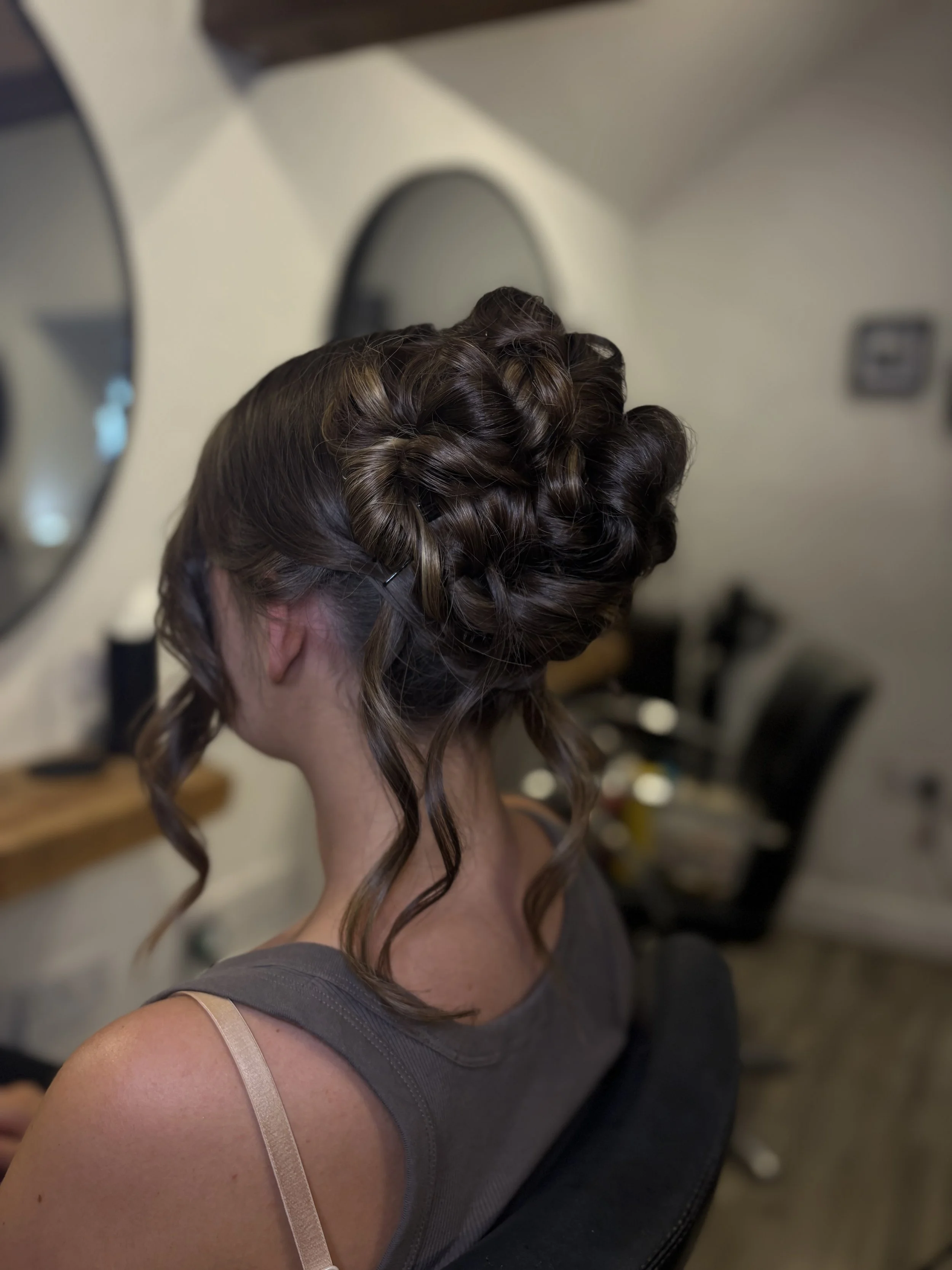 Back view of a woman with brown, curled hair sitting in a room, wearing a gray sleeveless top with a beige strap, in front of a mirror and a wall with a thermostat.