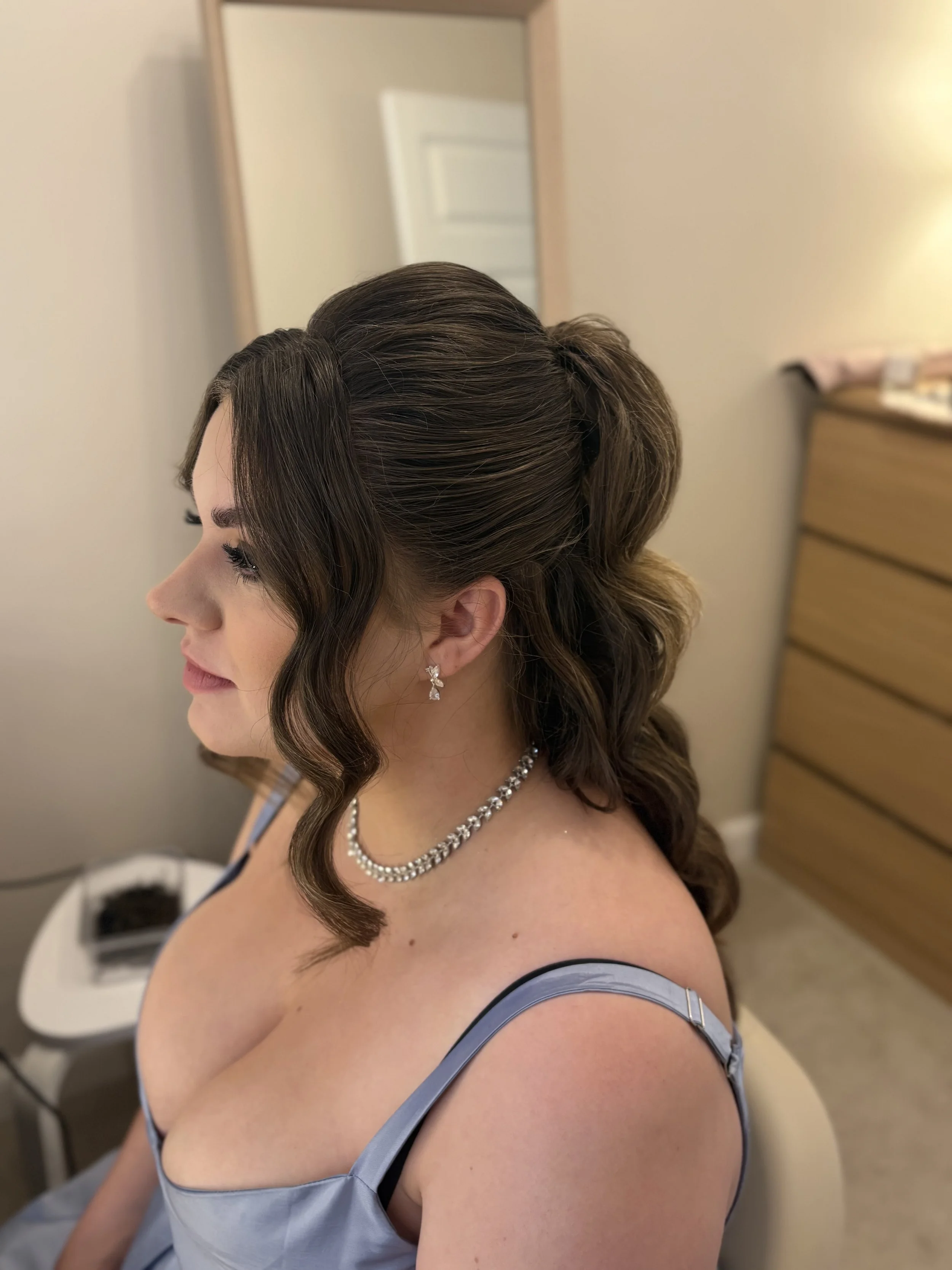 Profile of a young woman with brown, styled hair, dressed in a light blue dress, wearing diamond jewelry, in a room with a beige wall and a chest of drawers.