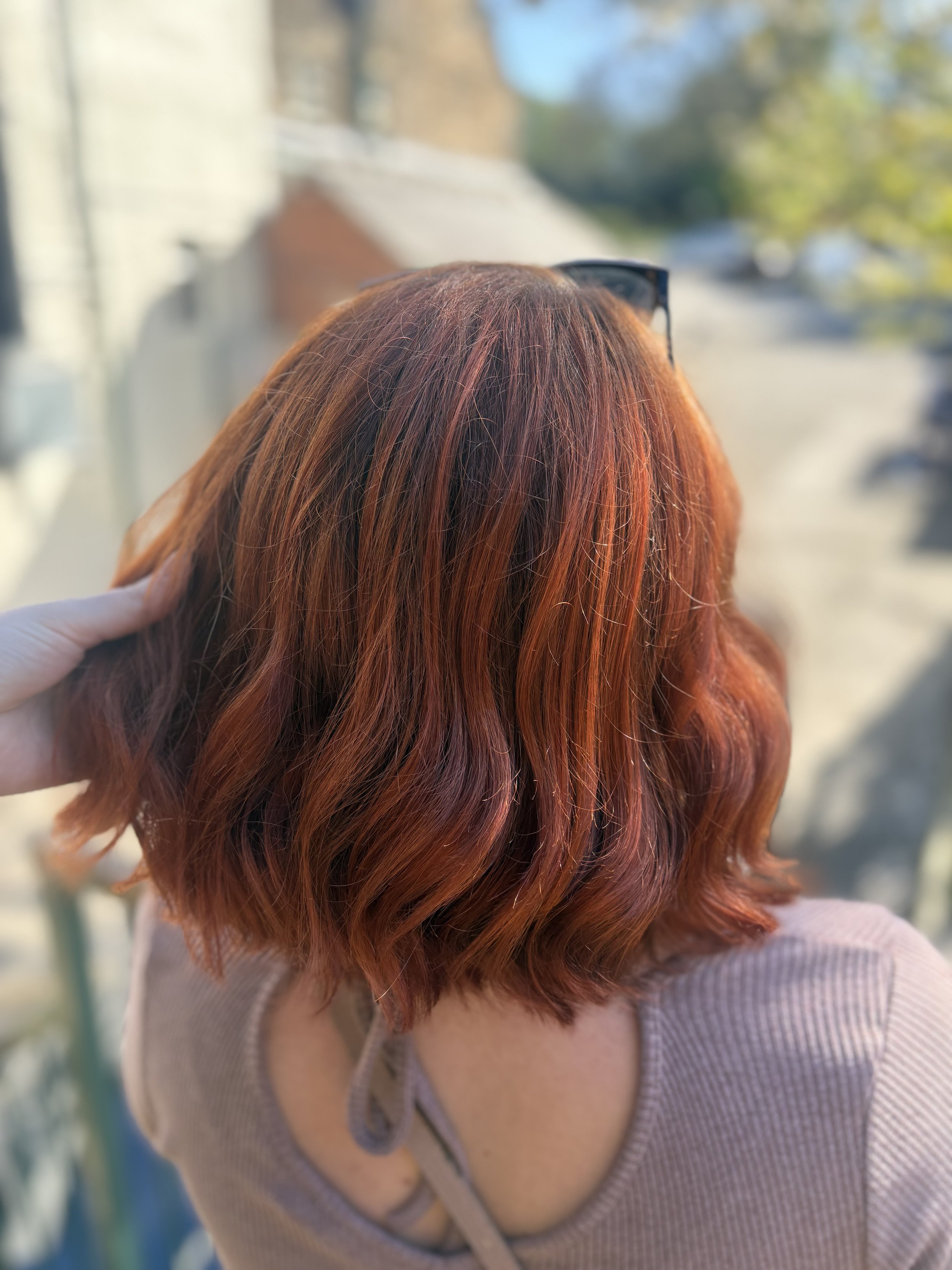 A woman with wavy, red hair wearing sunglasses on her head and a pink ribbed sweater, showing her back with her head turned slightly to the side outdoors.