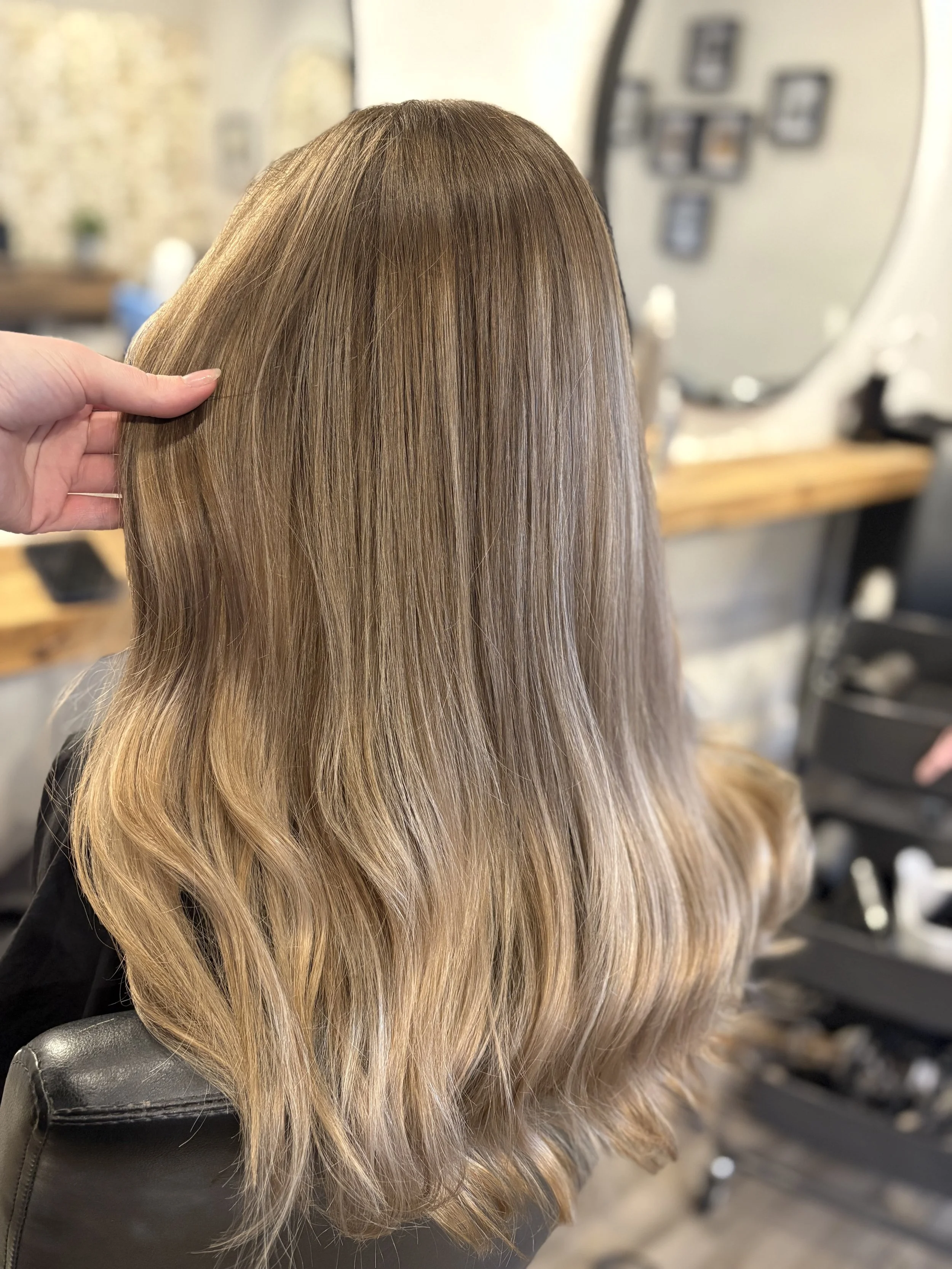 Close-up of a woman with long, shiny, ombre hair that transitions from light brown to blonde, sitting in a salon chair.