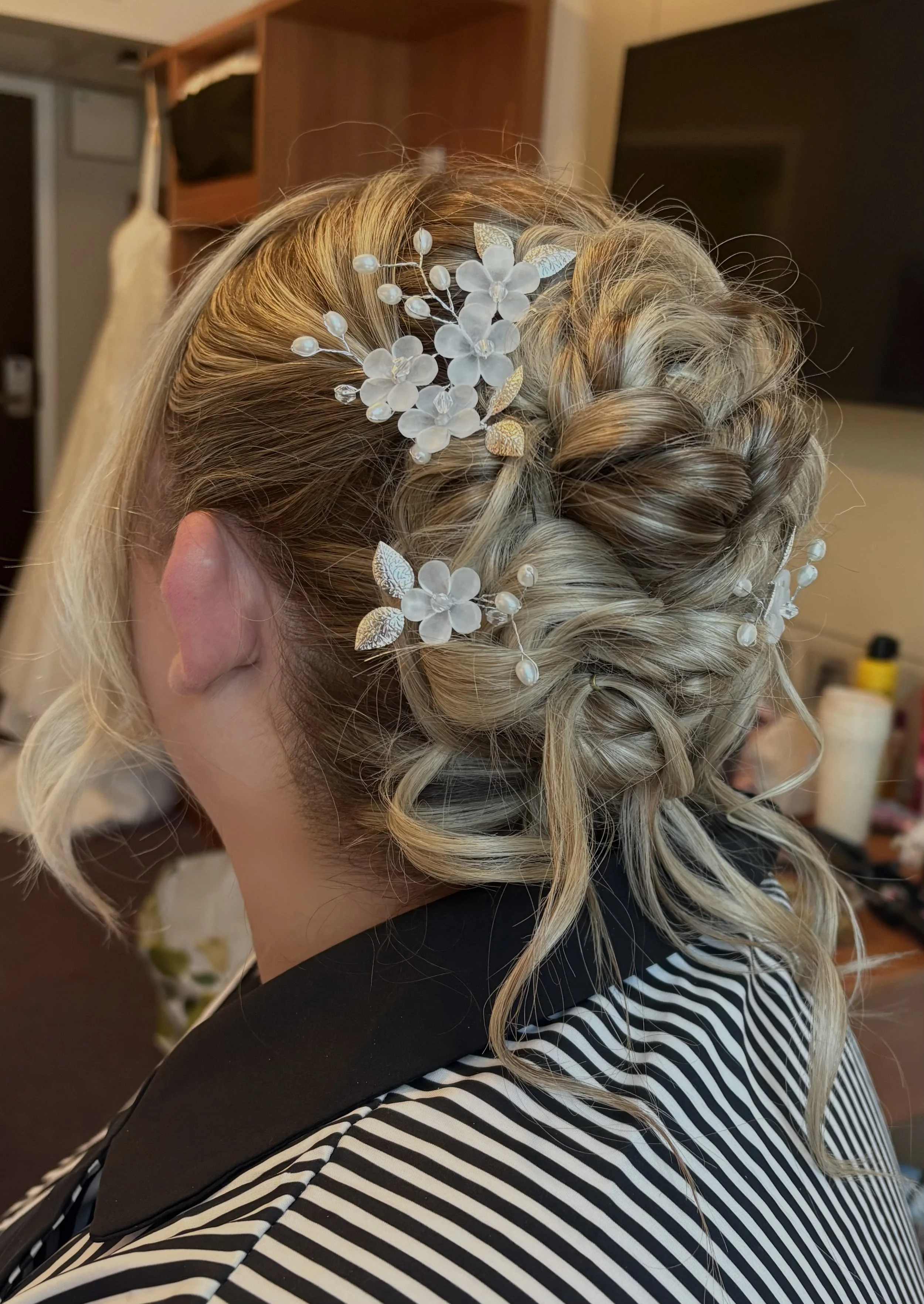 A woman with blonde hair styled in an updo adorned with a floral hair accessory, wearing a black and white striped garment, in a room with a TV and shelves in the background.