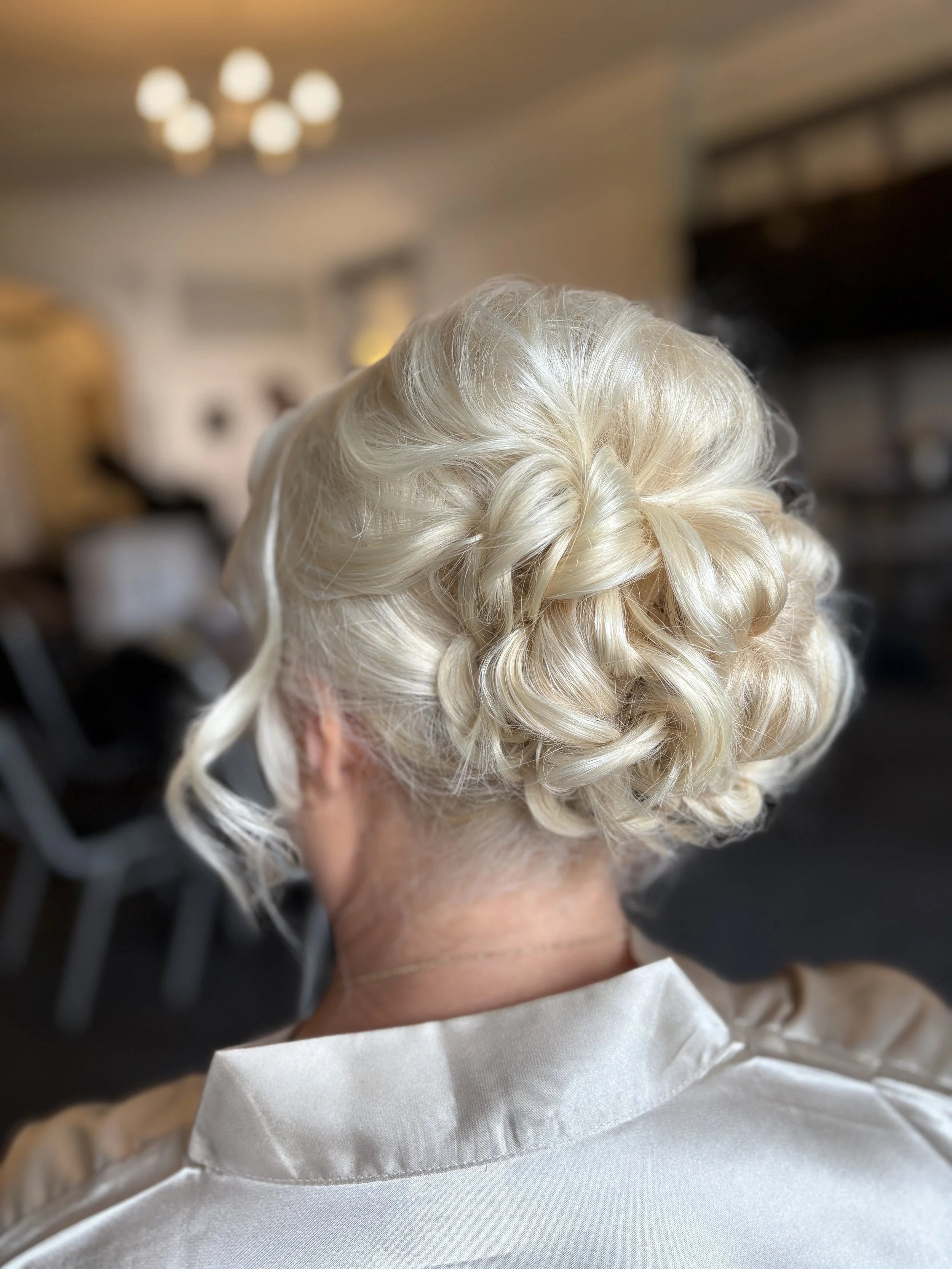 A hair stylist is styling a woman's platinum blonde hair with an intricate braid and curls in a salon.