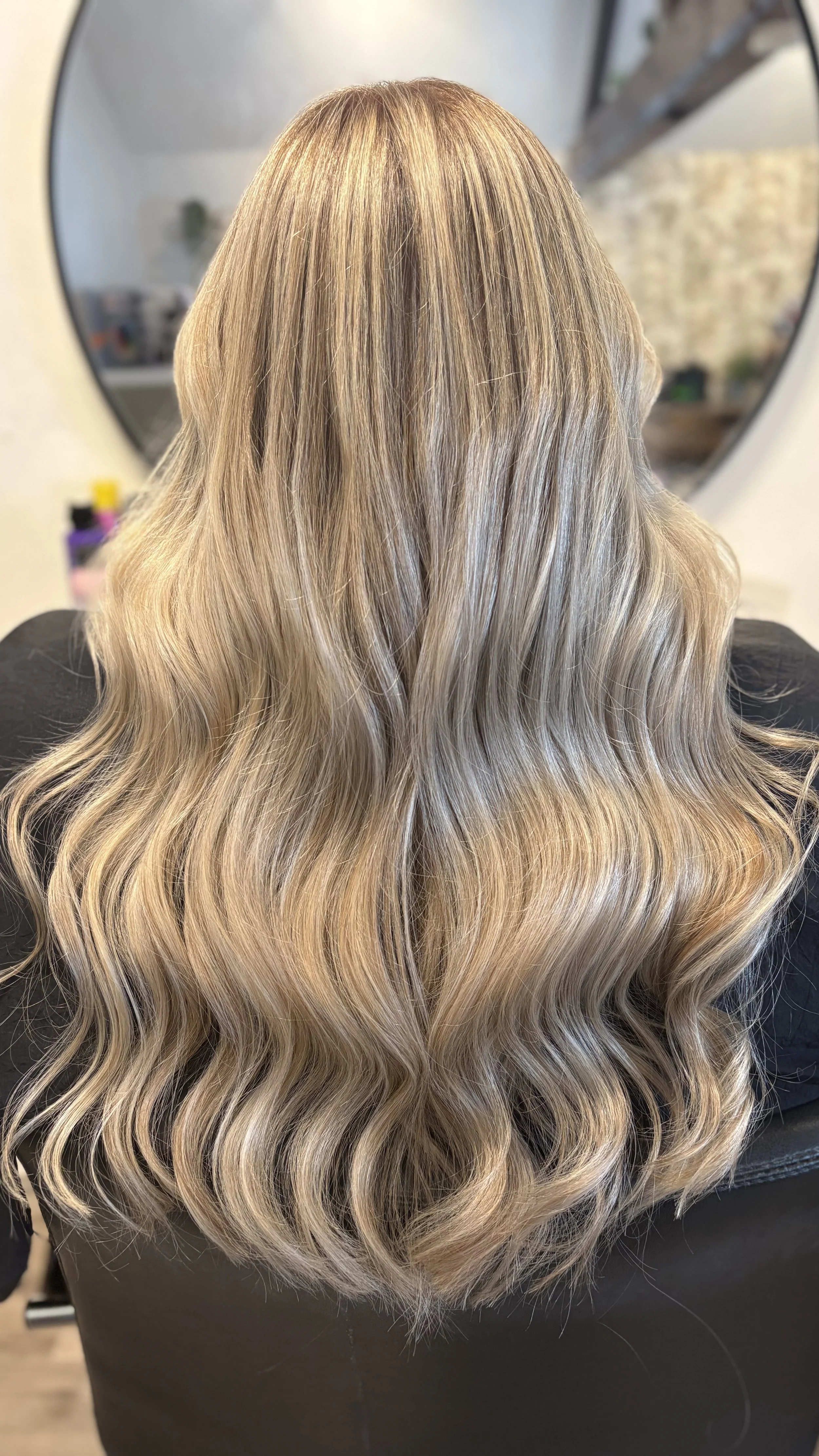 Back view of a person with long, wavy blonde hair with darker roots, sitting in front of a mirror.