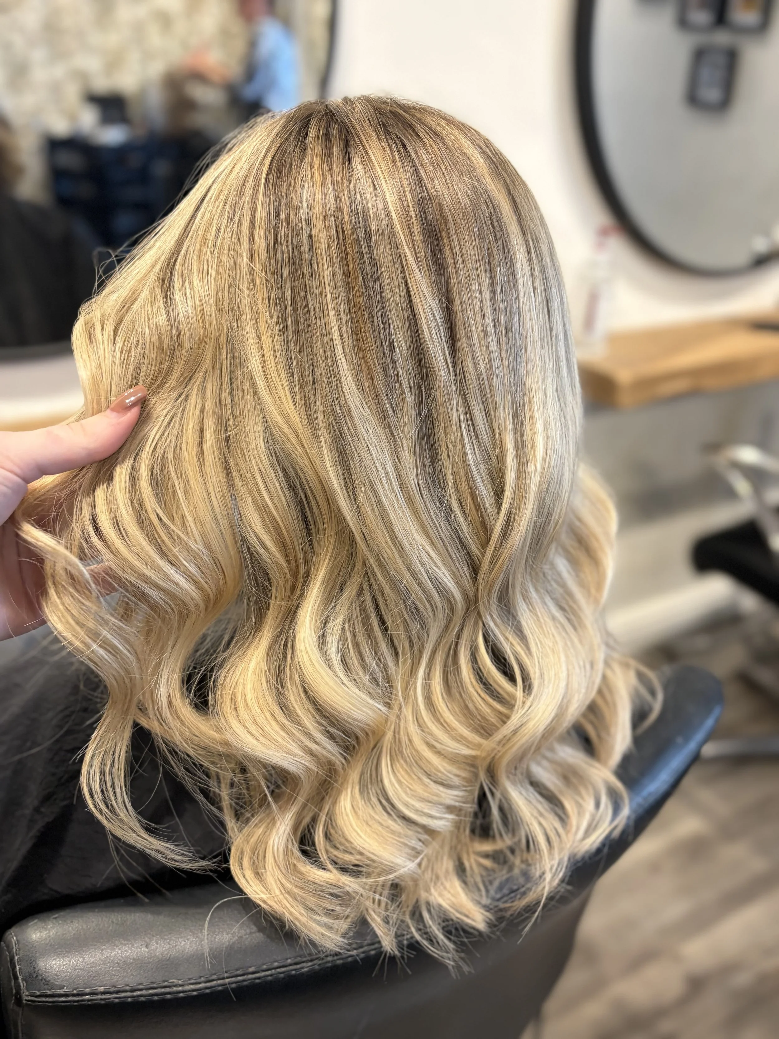 Close-up of a woman's blonde hair with loose curls, being held by someone’s hand, likely in a salon setting.