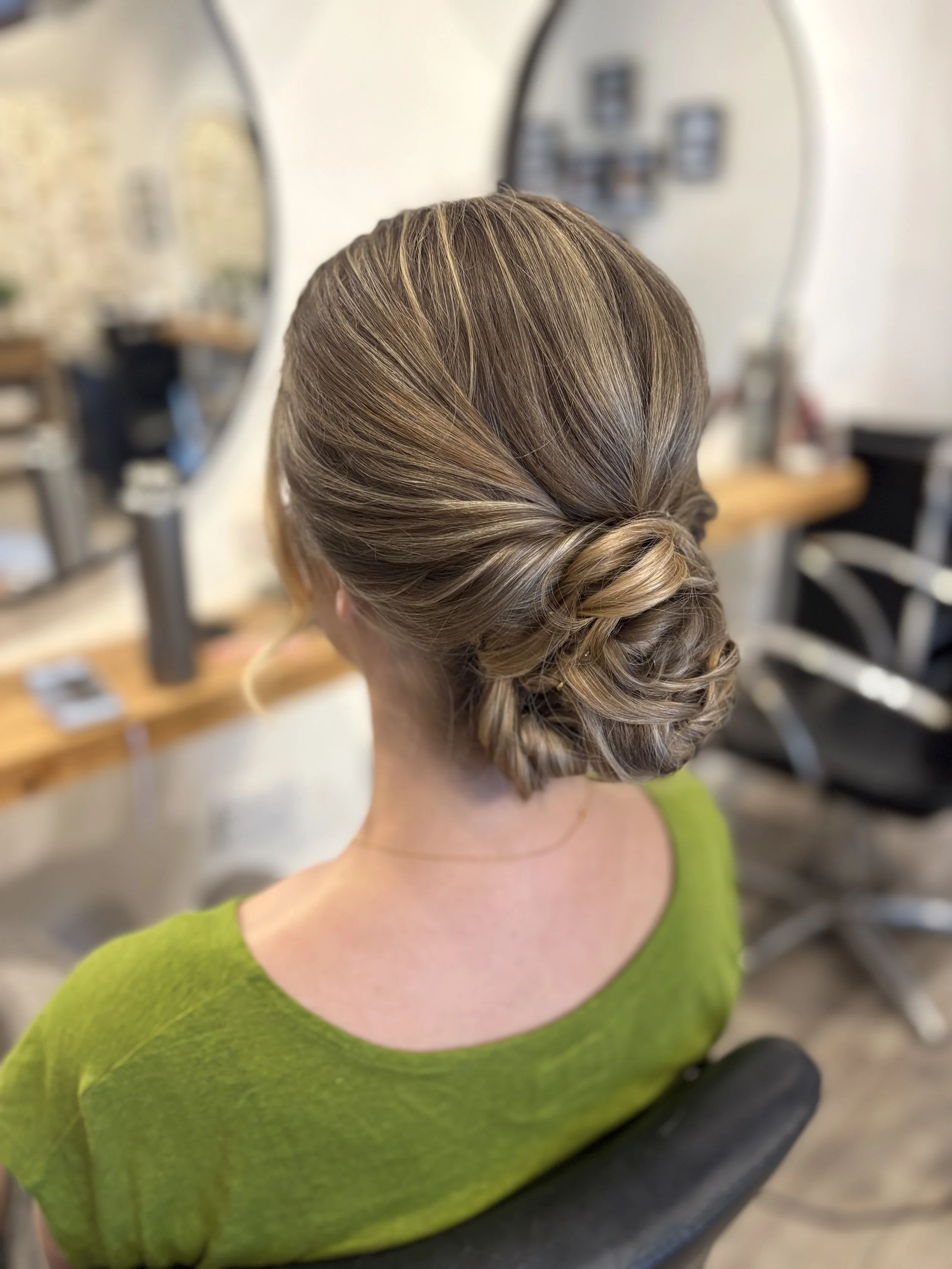 Back of a woman with styled, blonde hair in a loose updo, wearing a green top, sitting in a salon or studio with blurred objects and a mirror in the background.