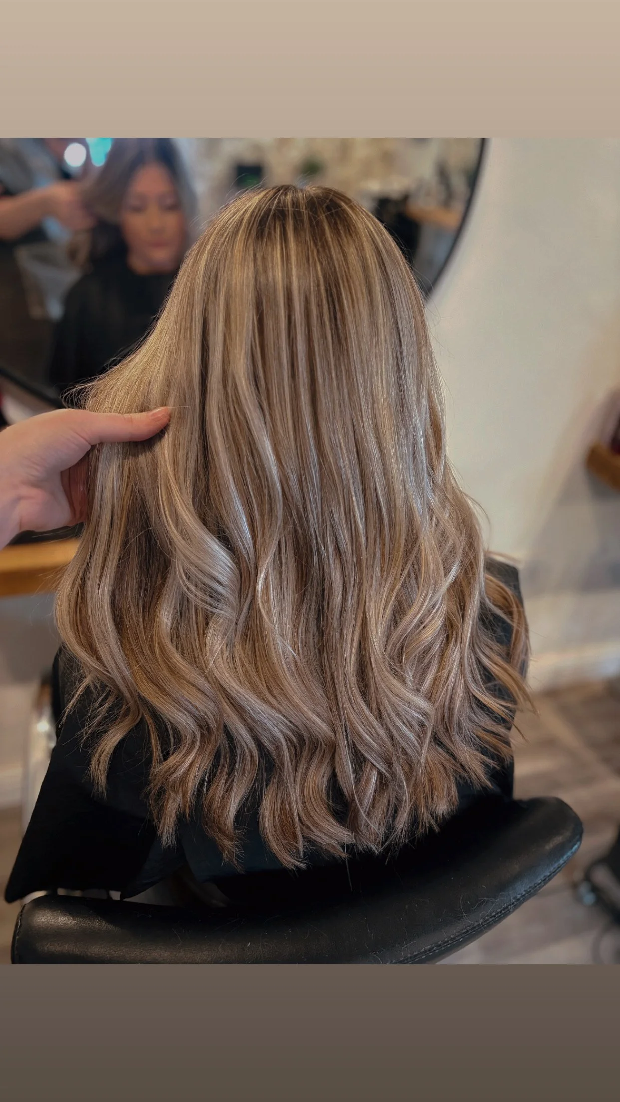 The image shows a woman with long, wavy blonde hair sitting in a salon chair, facing a mirror. A stylist is holding her hair, possibly styling or trimming it.