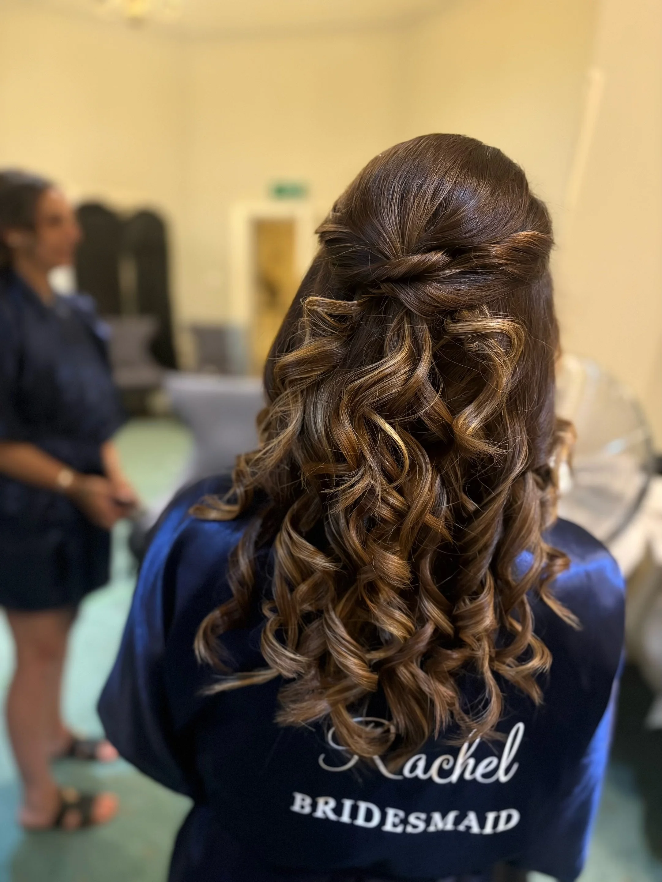 Back of a woman in a blue shirt with curling hair, wearing a vest with the words 'Rachel BRIDESMAID' on it.