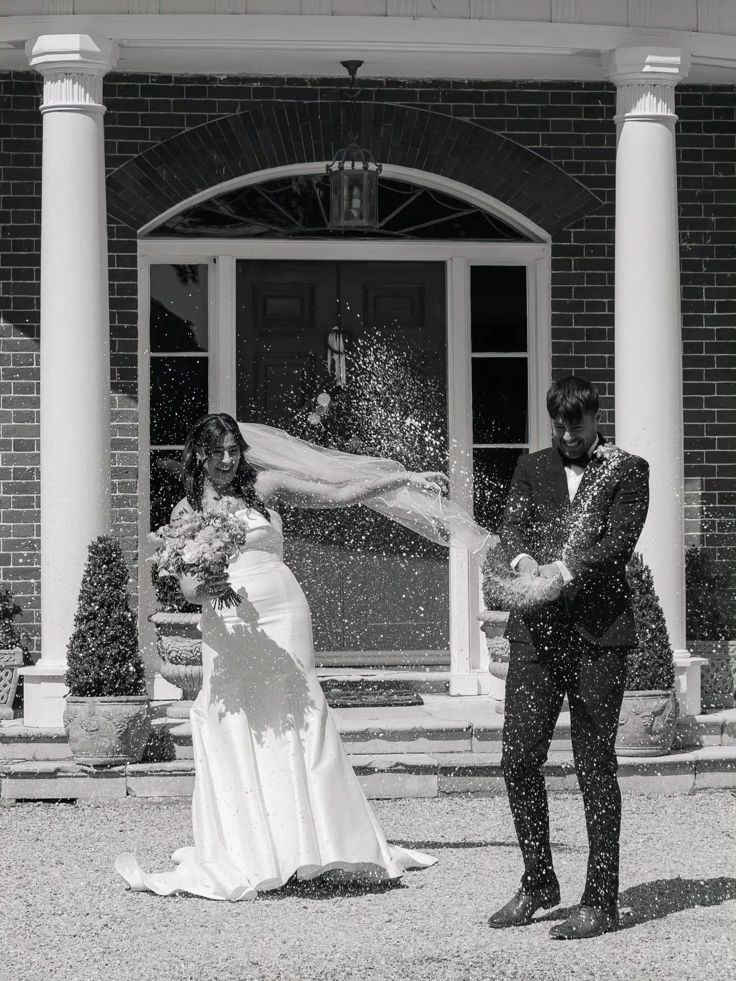 A bride and groom celebrating outside a brick house with columns, as they are showered with rice or confetti.