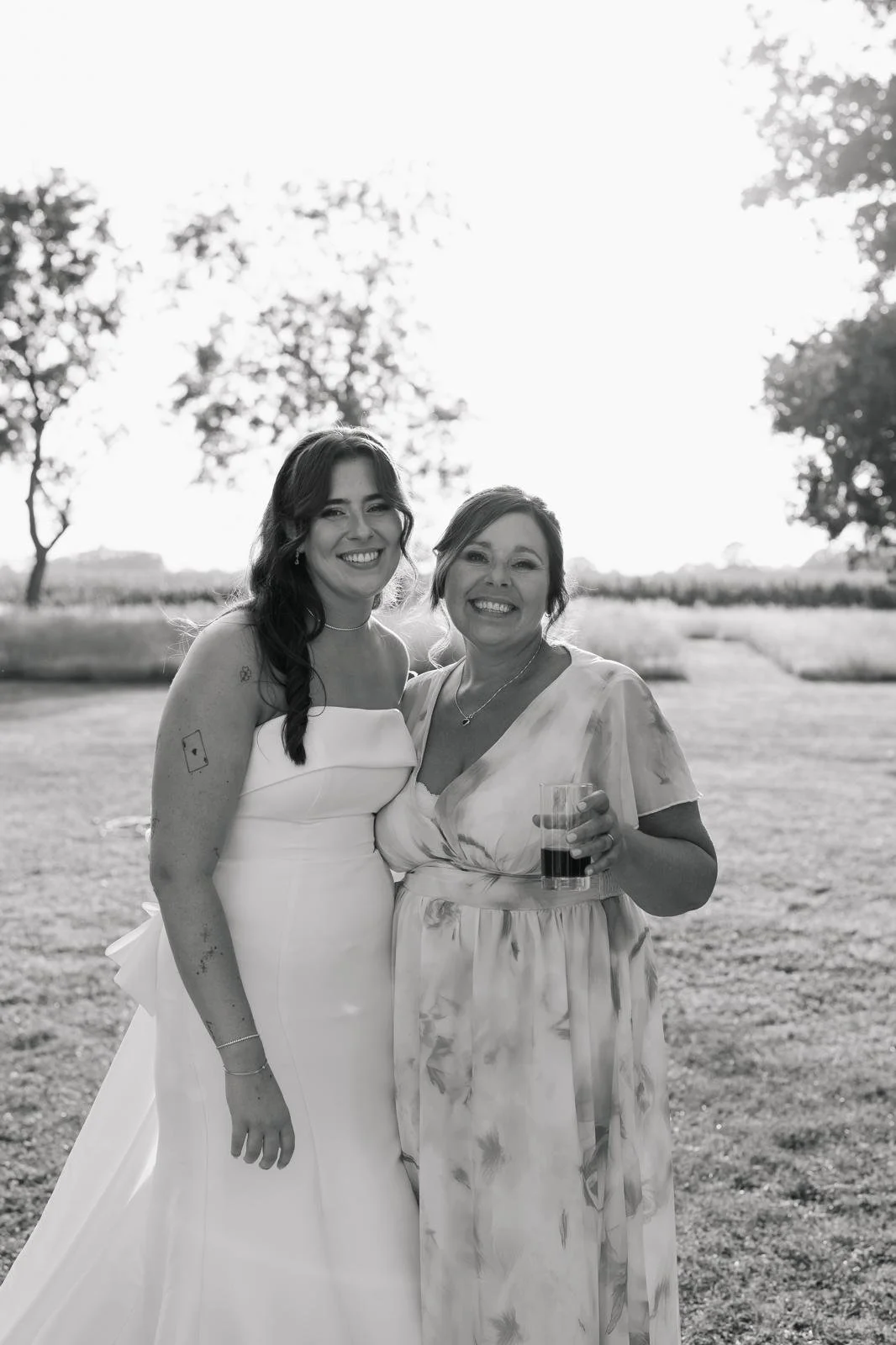 Two women smiling outdoors in a park, one in a wedding dress and the other holding a drink