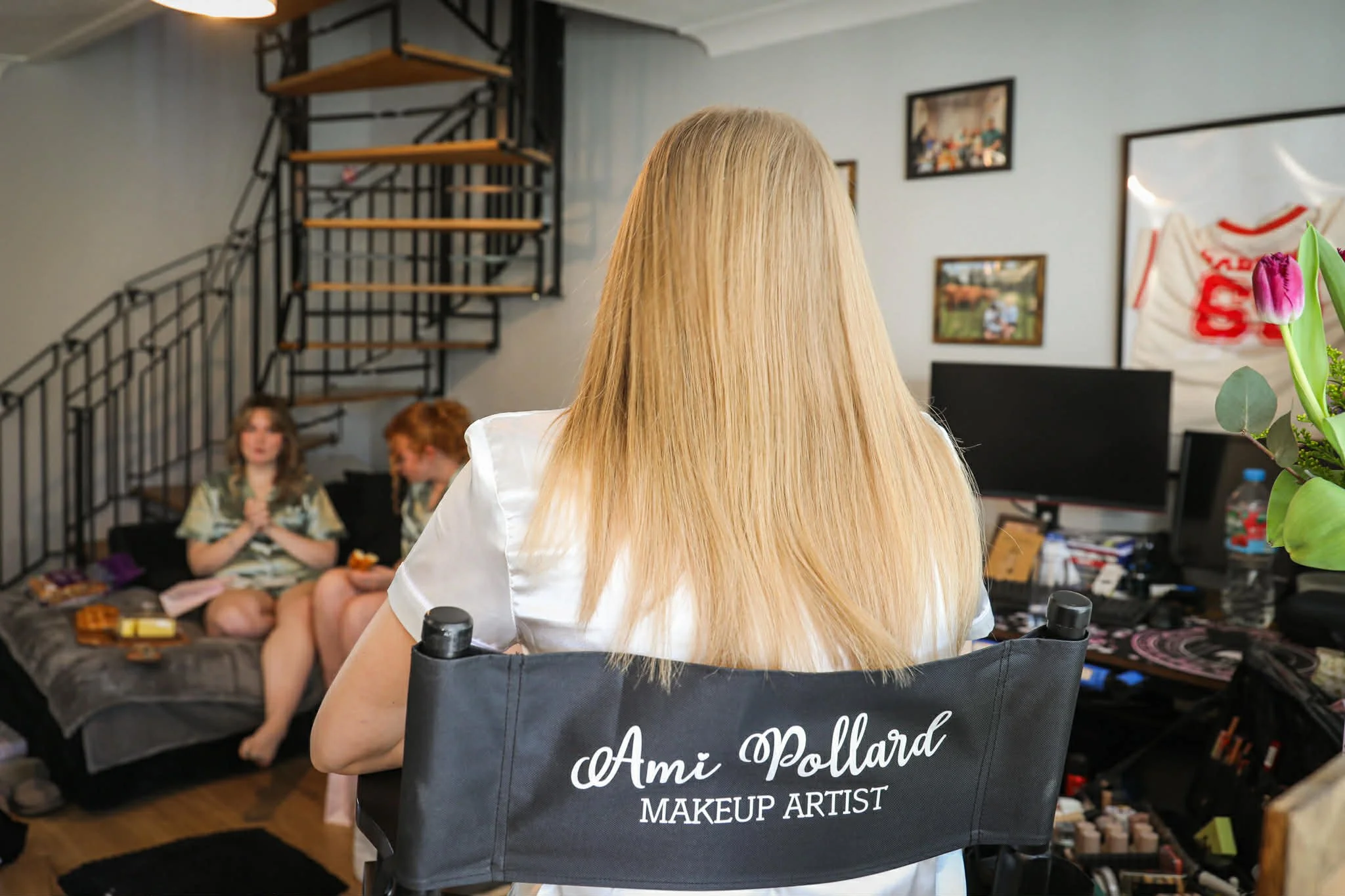 A woman in a salon chair with her back to the camera, that says 'Ami Pollard Makeup Artist'. She has long blonde hair. In the background, two women are sitting on a couch, one with long brown hair and the other with short red hair, both engaged in conversation. The salon has framed pictures on the wall, some shelves, and a large bag with red lettering.