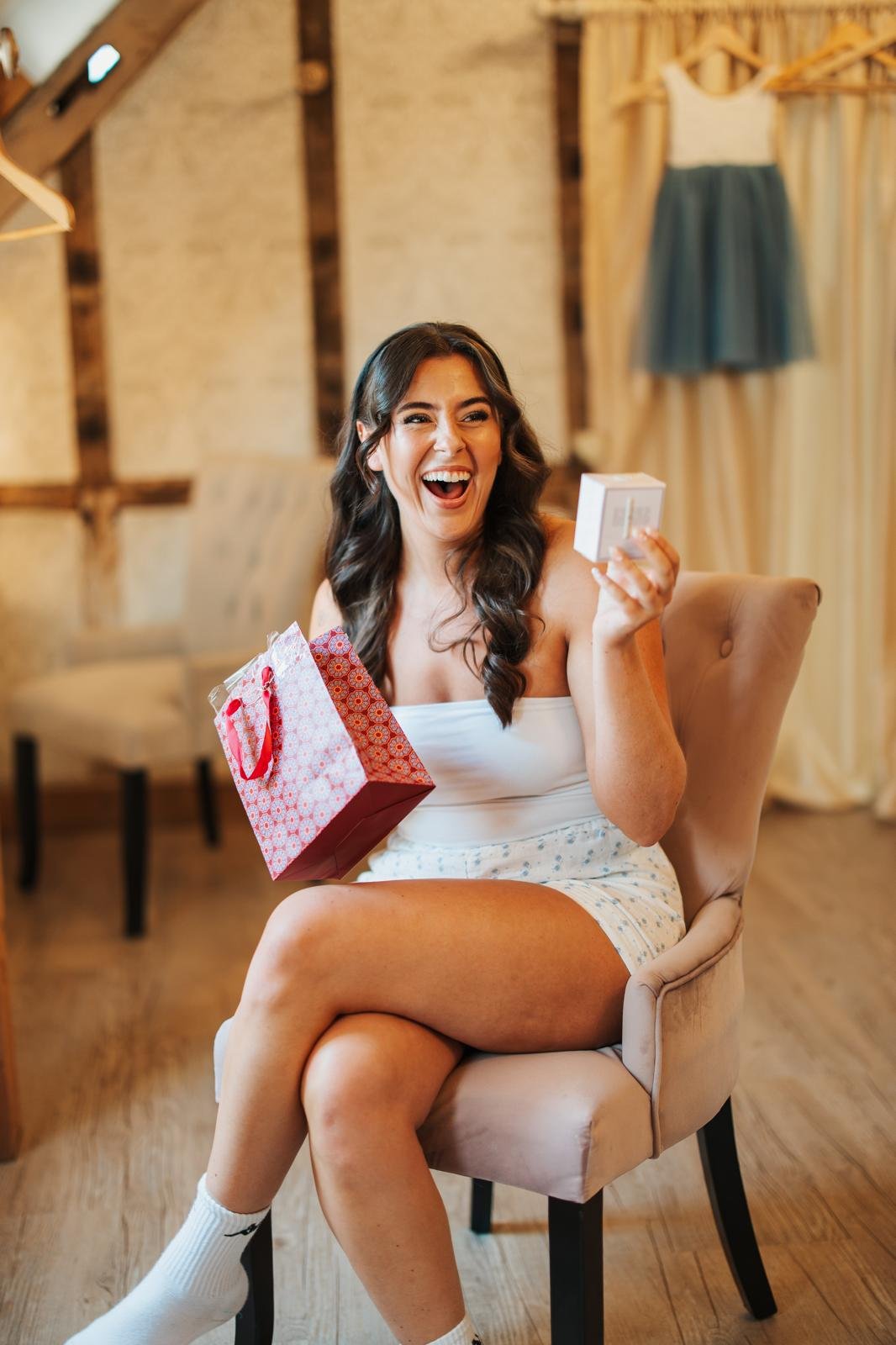 A woman with long dark hair, wearing a white tank top and shorts, sitting on a beige chair, smiling and holding a small gift box in one hand and a pink gift bag in her lap, in a rustic room with wood accents and dresses hanging in the background.