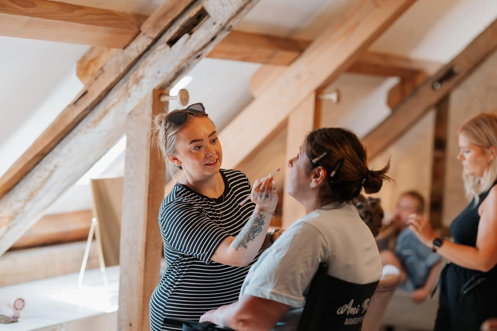 A woman with blond hair, wearing a black and white striped top, is applying makeup or face paint to a seated woman with dark hair in an indoor setting with wooden beams and other women in the background.