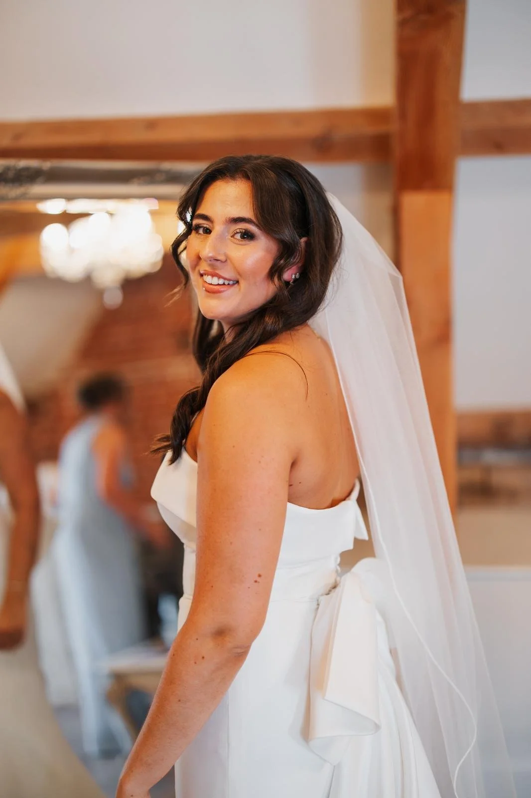 A smiling bride with dark brown wavy hair in a white wedding dress and veil, indoors with wooden beams and a chandelier in the background.
