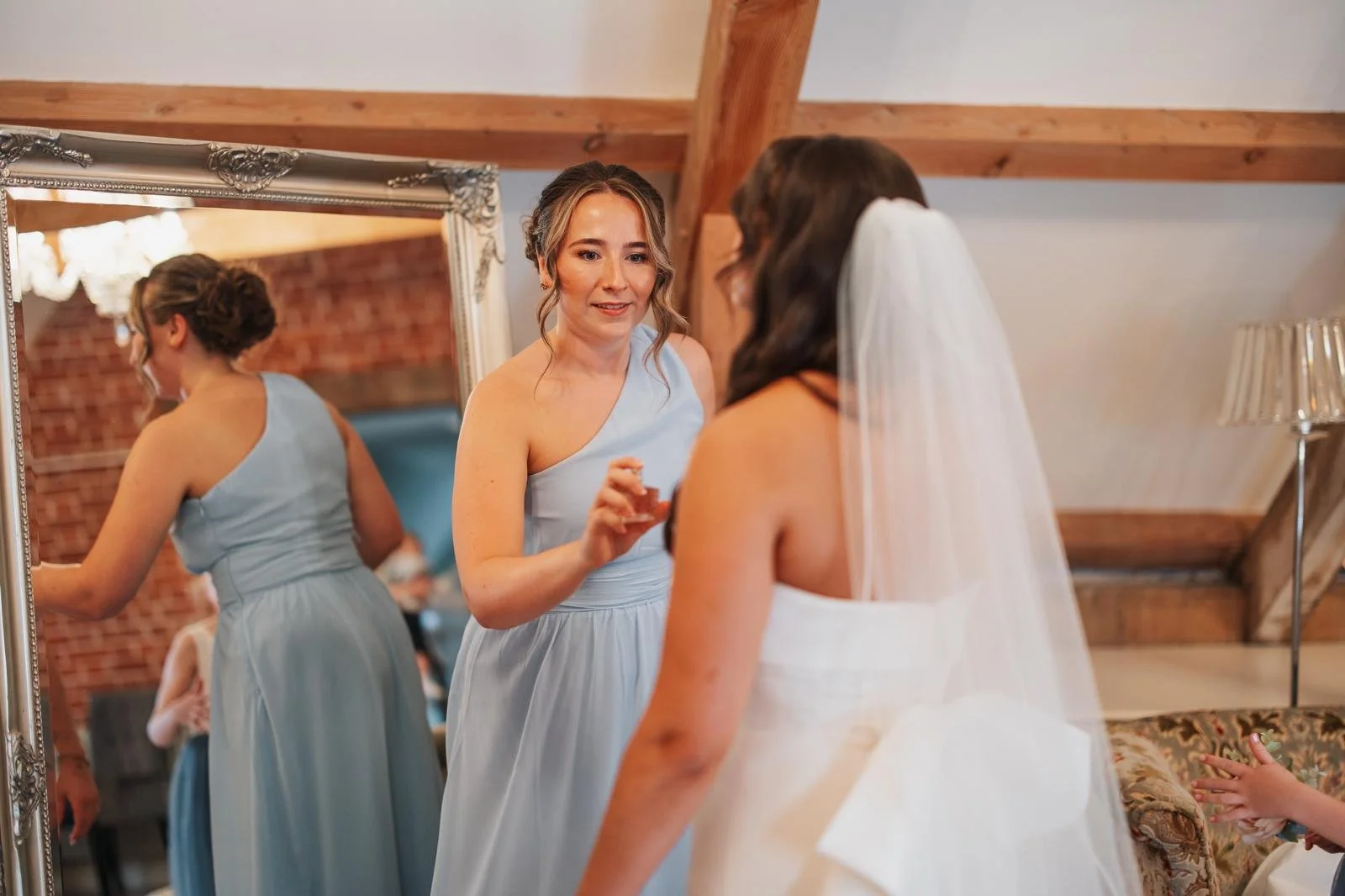 Bridal preparation with women getting ready, one in a white wedding dress and veil, another in a light blue bridesmaid dress, standing near a full-length mirror in a rustic, wooden-beamed room.