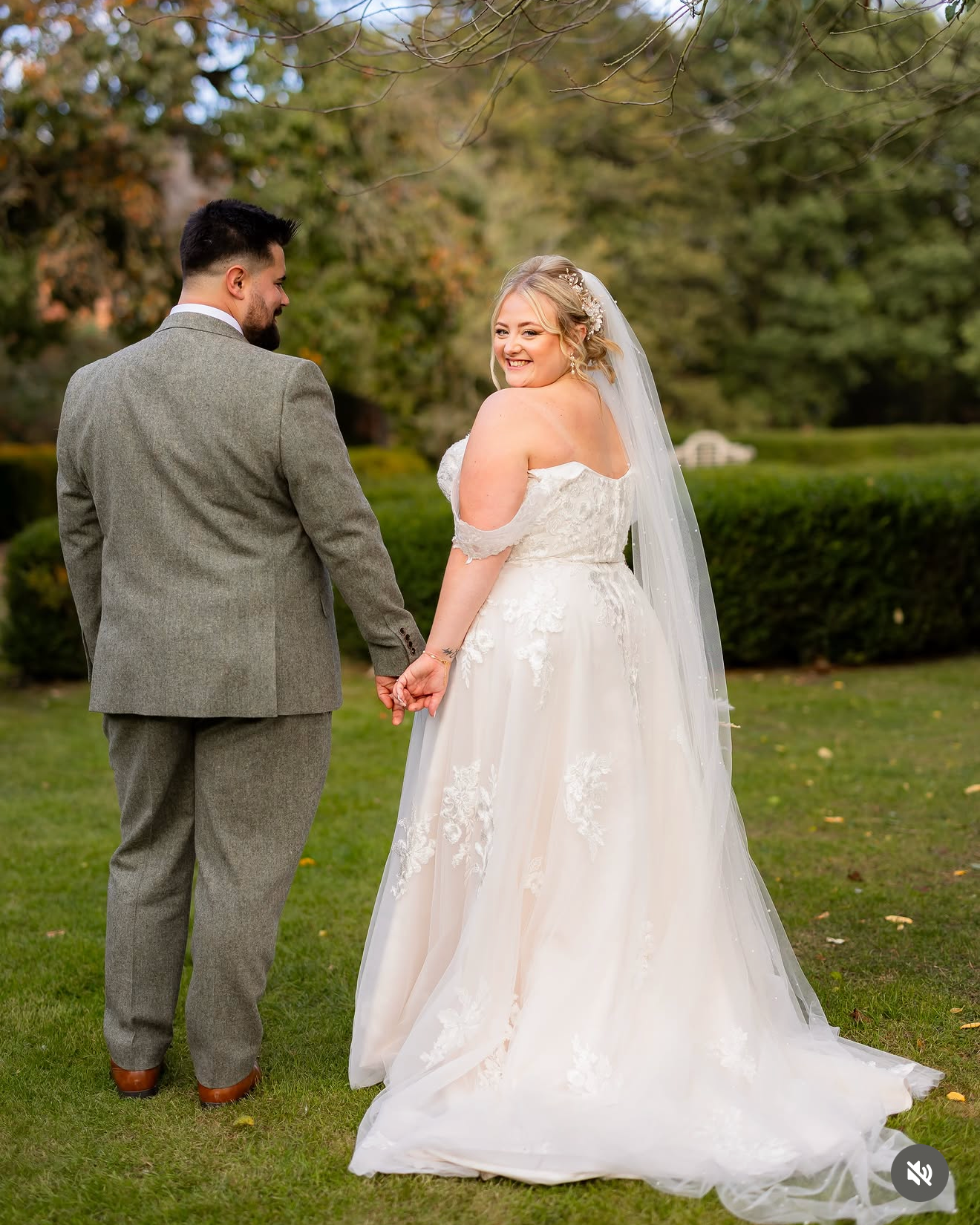 A newlywed couple holding hands outdoors on a grassy area with trees in the background, smiling at each other, dressed in wedding attire. The bride wears a white off-the-shoulder wedding gown with lace details and a veil, and the groom wears a gray suit.
