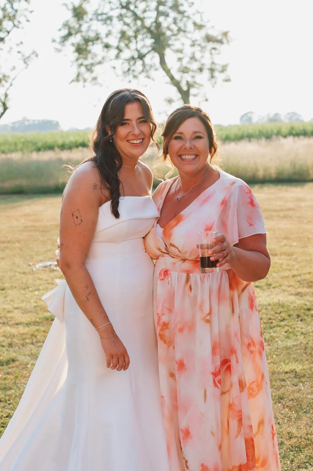 Two women smiling outdoors at a wedding, one in a white wedding dress and the other in a floral dress holding a glass of dark beverage.