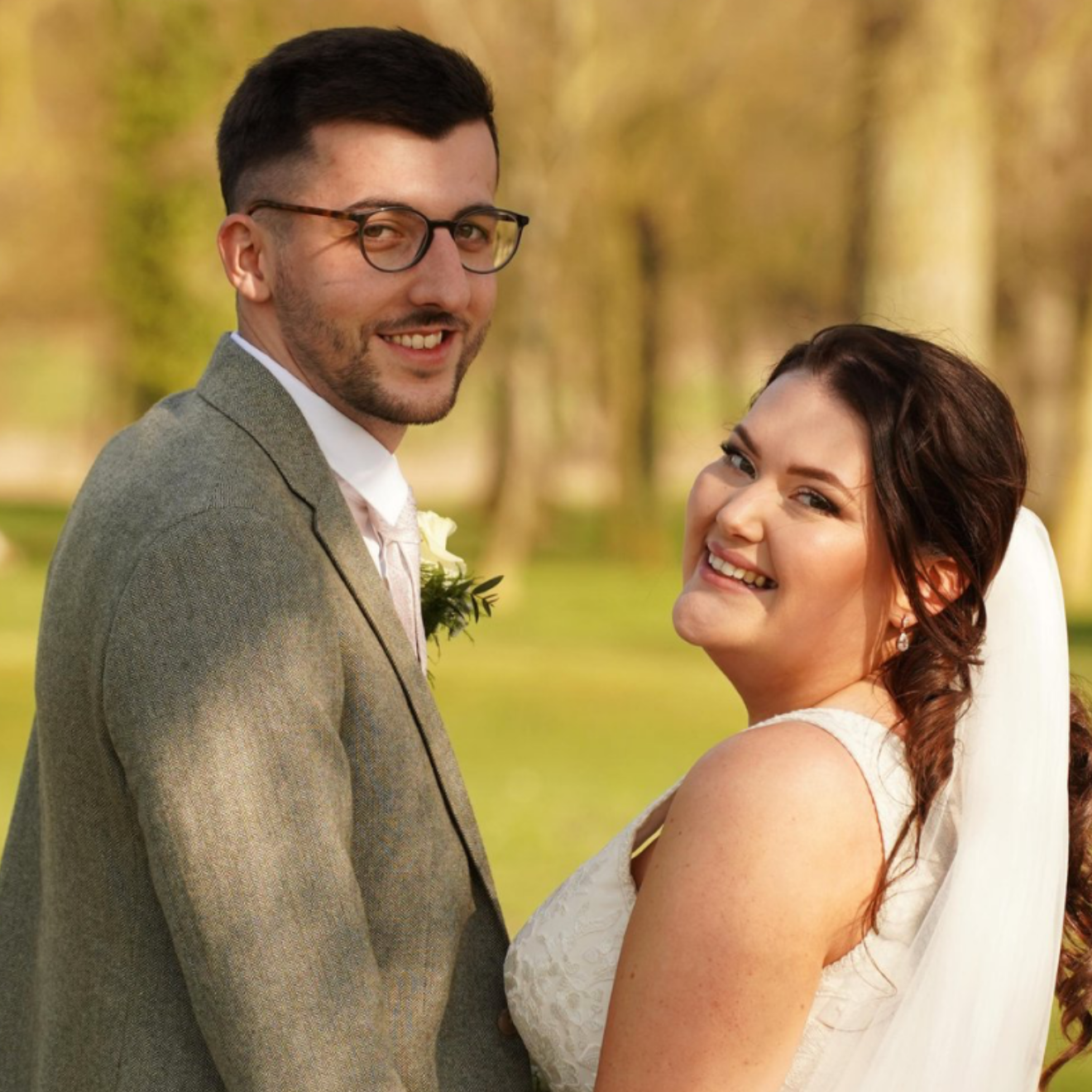 A newlywed couple smiling outdoors during daytime, with trees in the background. The groom wears glasses, a suit, and a boutonniere, and the bride is in a white wedding dress with a veil.