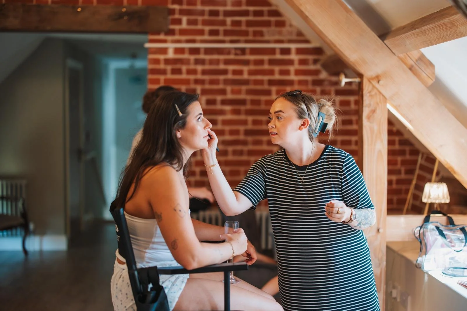 A makeup artist applies makeup to a woman with long dark hair, tattoos, and wearing a white tank top, in a room with exposed brick walls and wooden beams. The woman is seated at a makeup station with makeup brushes and tools on the counter.