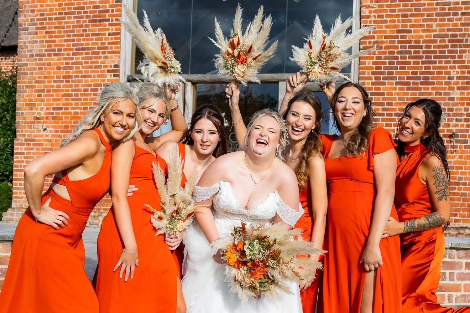 Group of women celebrating at a wedding, wearing matching bright orange dresses with the bride in a white gown holding a bouquet of flowers, standing in front of a brick building.