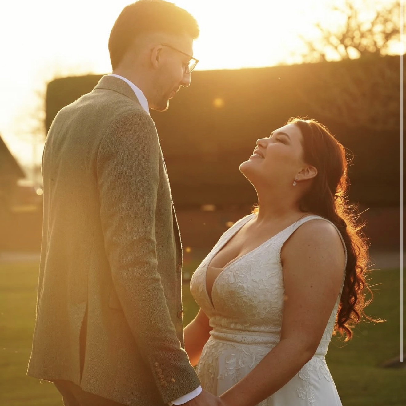 A couple standing outdoors during sunset, gazing into each other's eyes, holding hands. The man is wearing a tan suit with glasses, and the woman is in a white lace dress with long red hair.