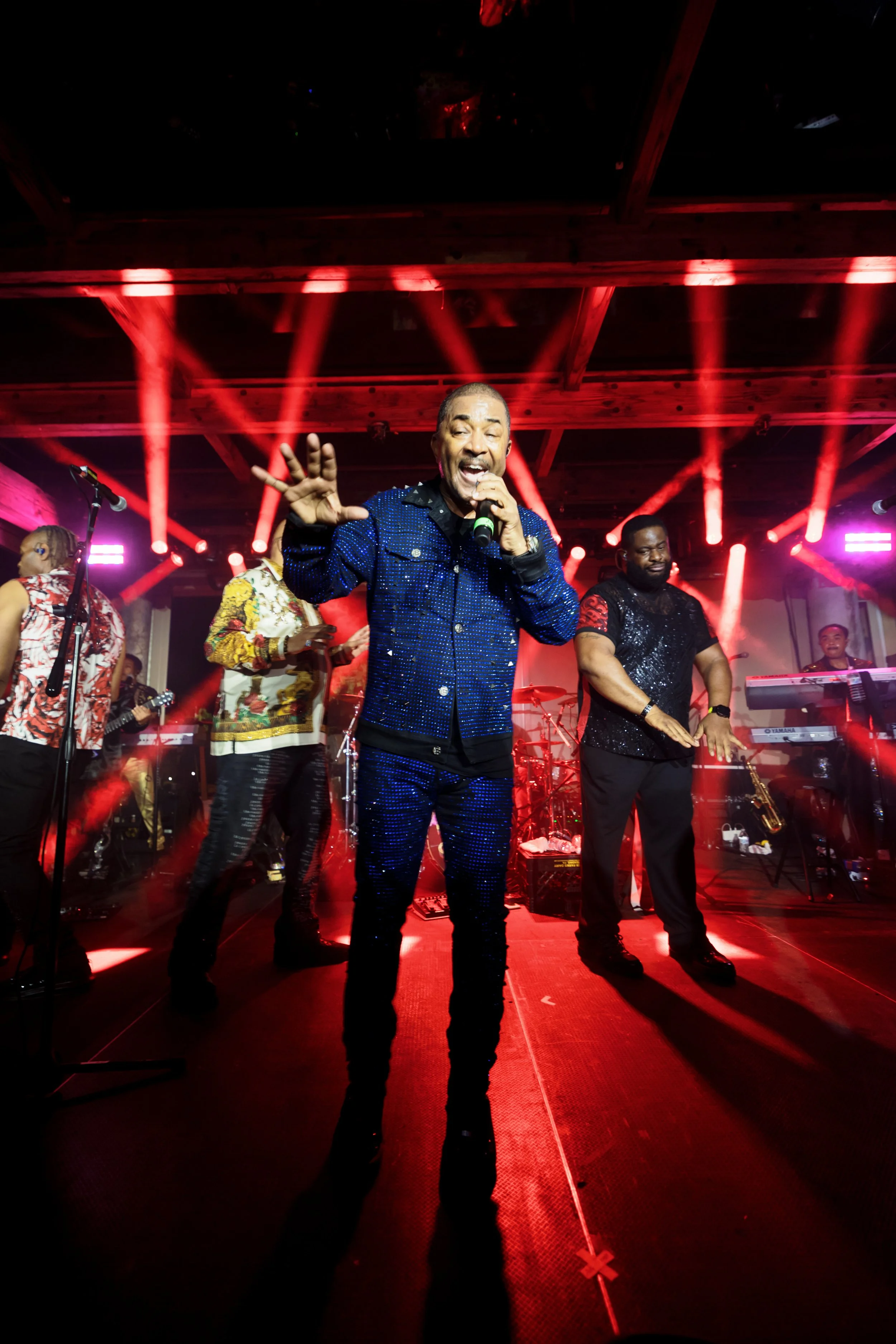 Singer performing into a microphone with a band on a concert stage illuminated by red lights