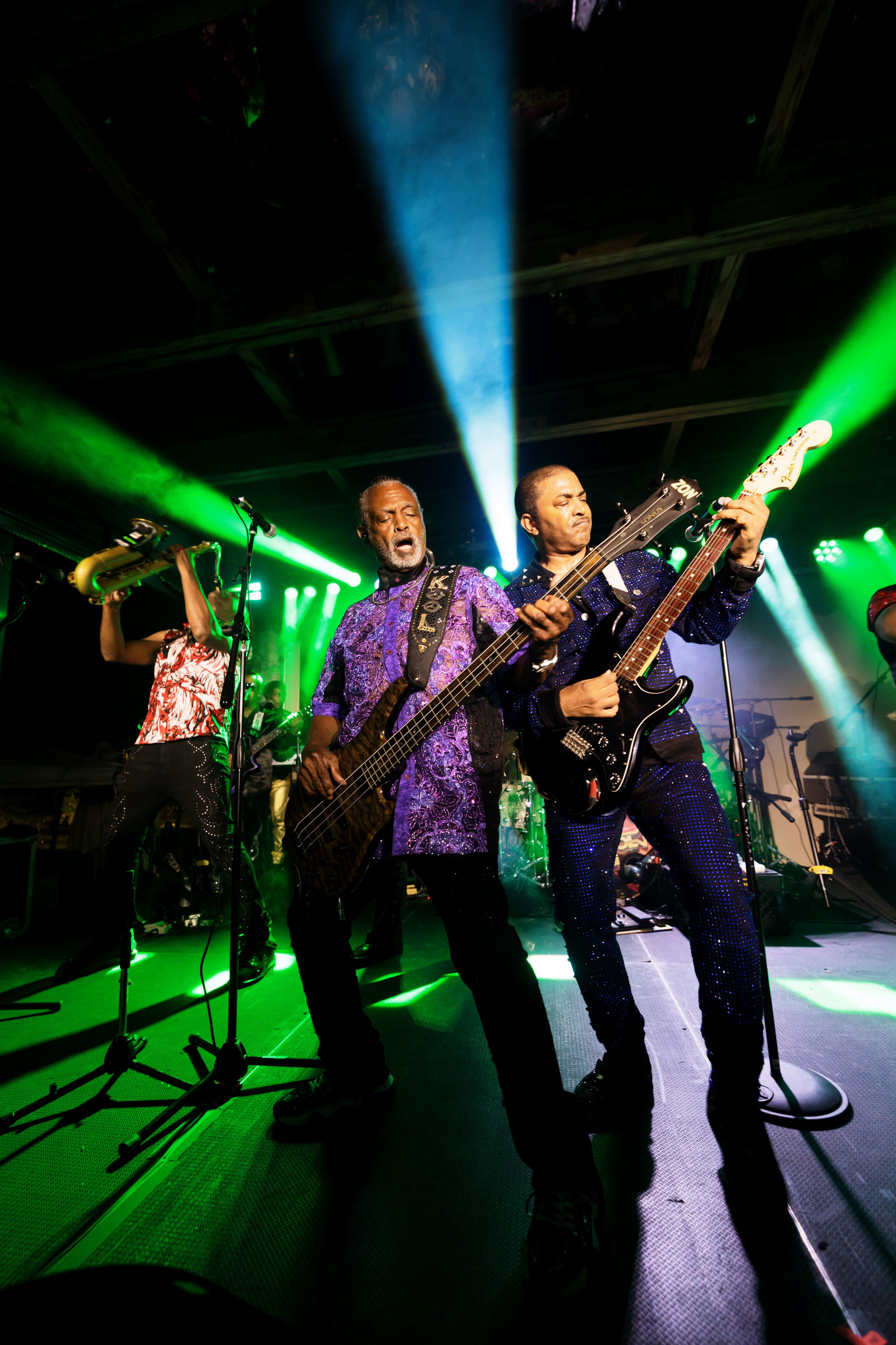 Live band performing onstage under green and blue lights, featuring a bassist, guitarist, and saxophonist.
