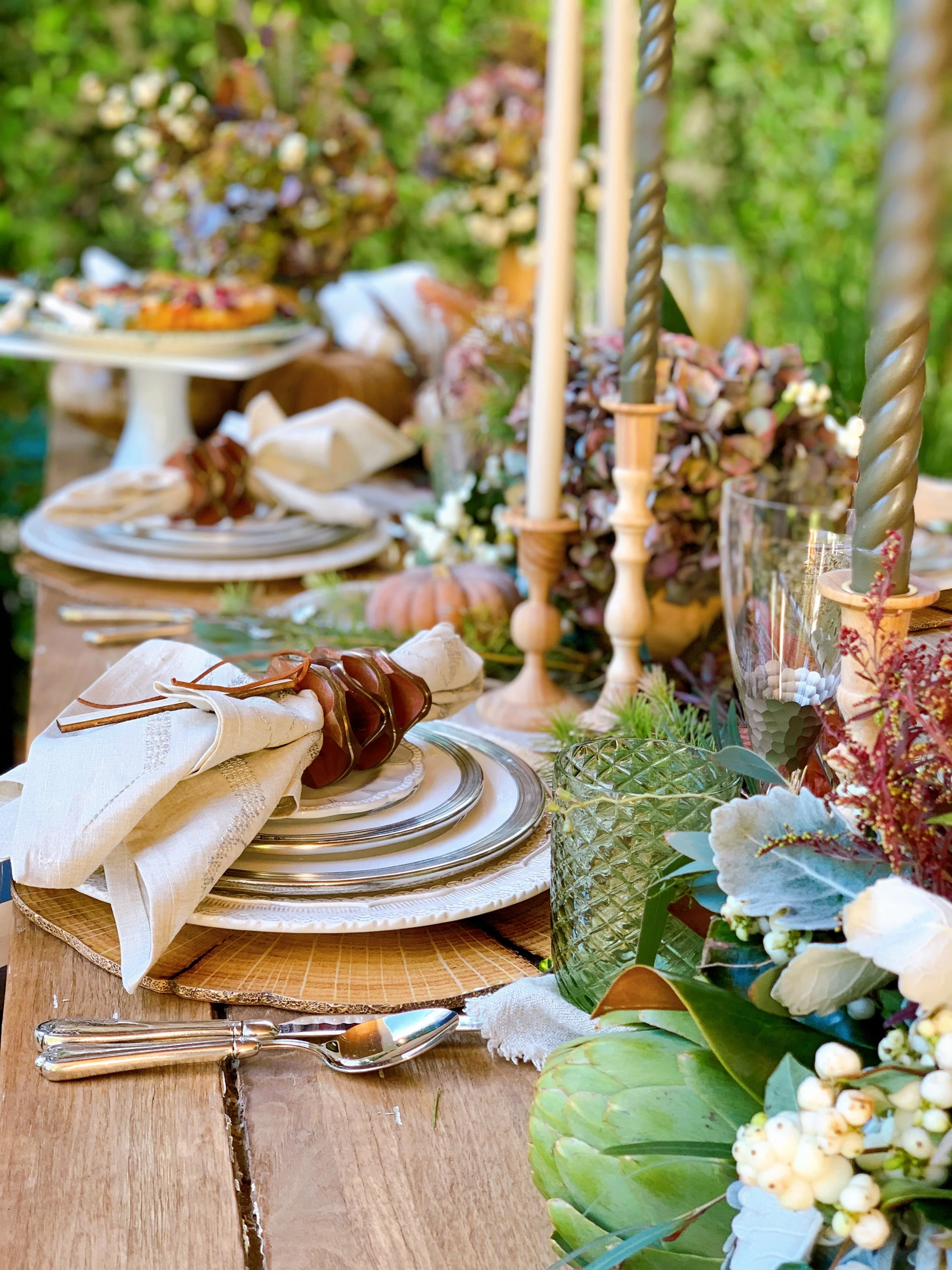 Elegant outdoor table setting with stacked plates, folded napkins, silverware, candles, glassware, pumpkins, and lush floral greenery