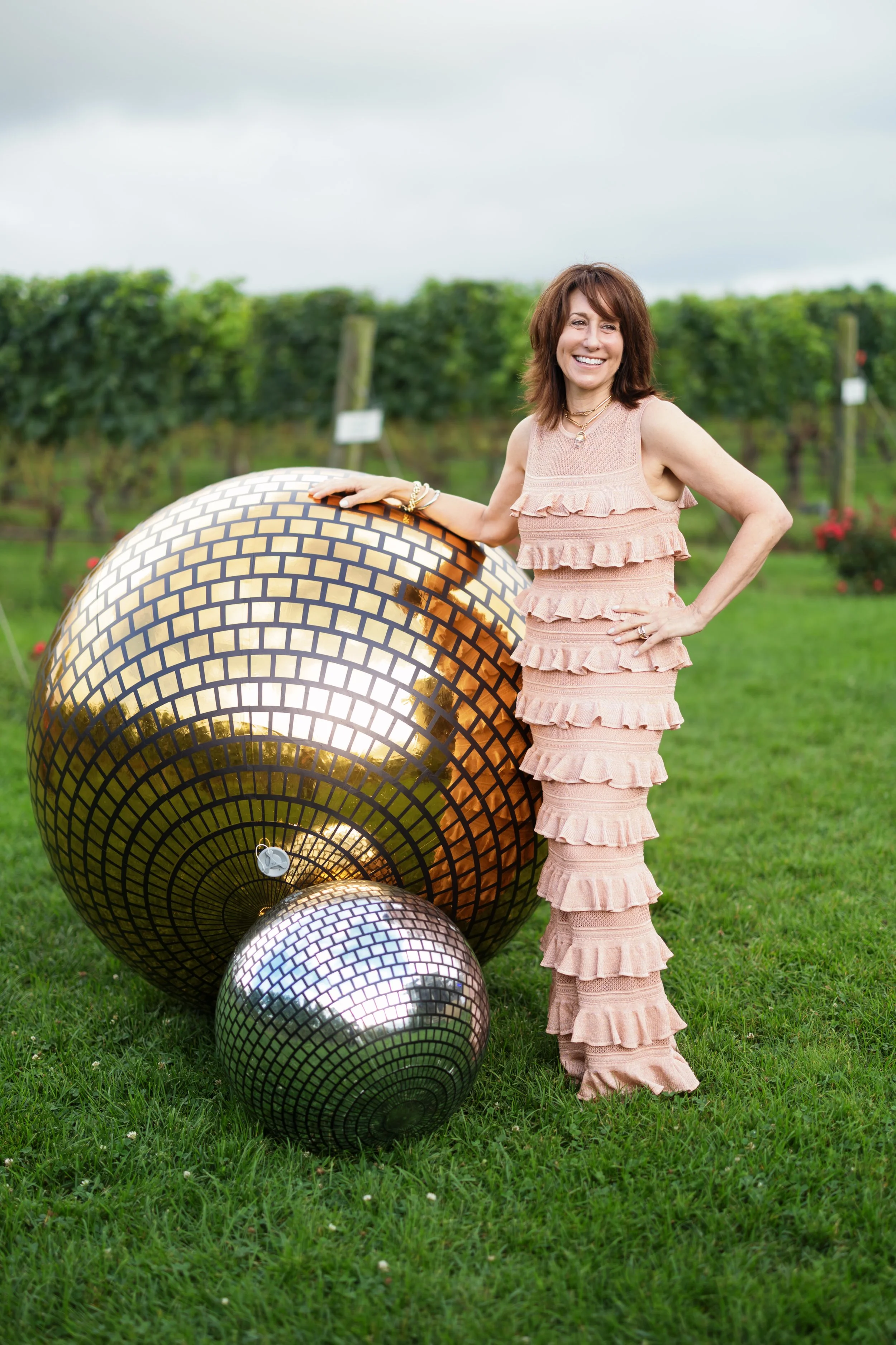 Smiling woman in a pink ruffled dress standing beside large mirrored mosaic spheres in a garden