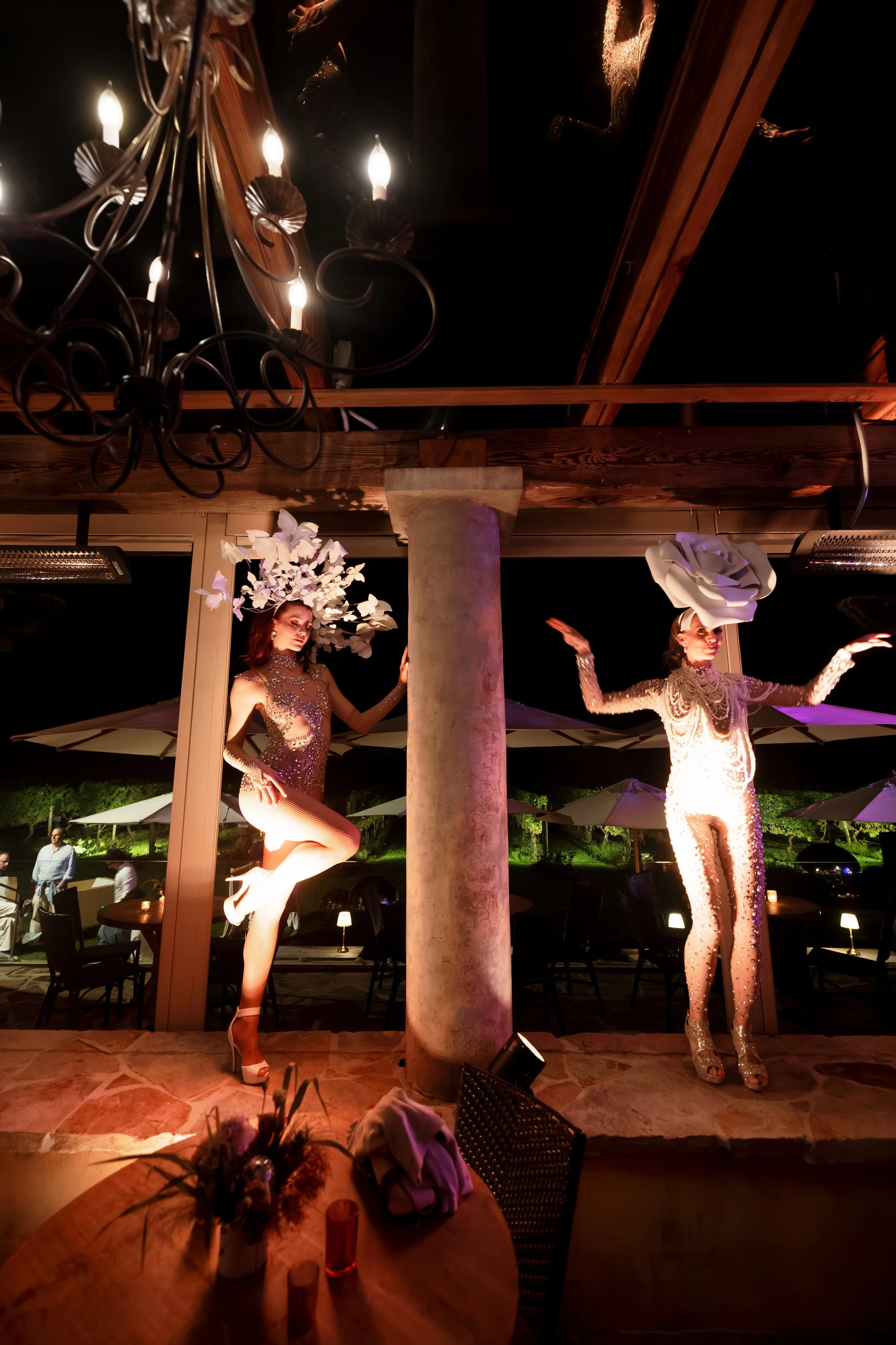 Two costumed dancers perform a glamorous nighttime stage show under chandeliers and spotlights at an outdoor venue.