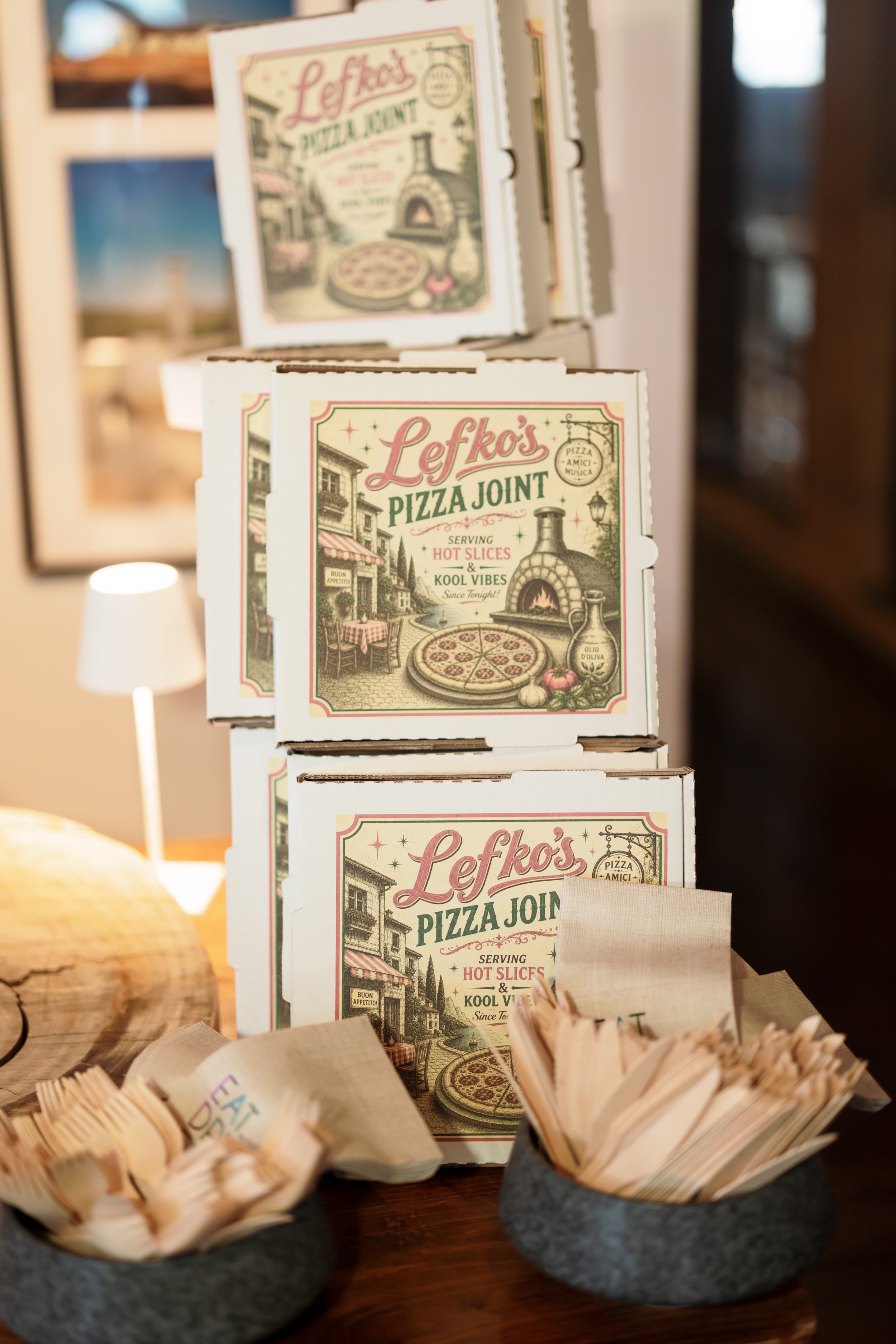Italian-style pizza boxes labeled Lefko’s Pizza Joint, with baskets of wooden utensils on a restaurant counter.