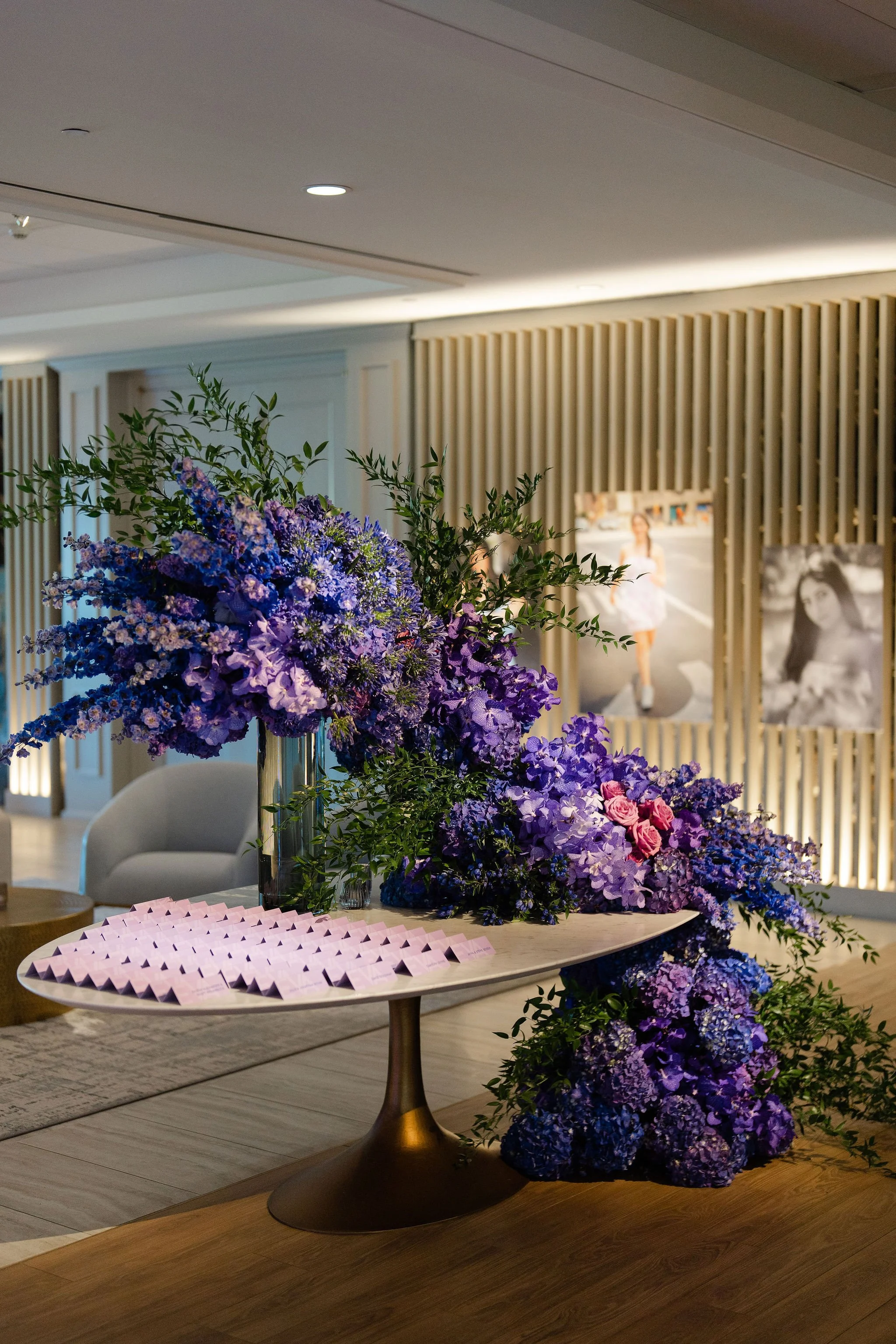 Elegant interior display featuring large purple and blue floral arrangements, pink place cards, and framed artwork.