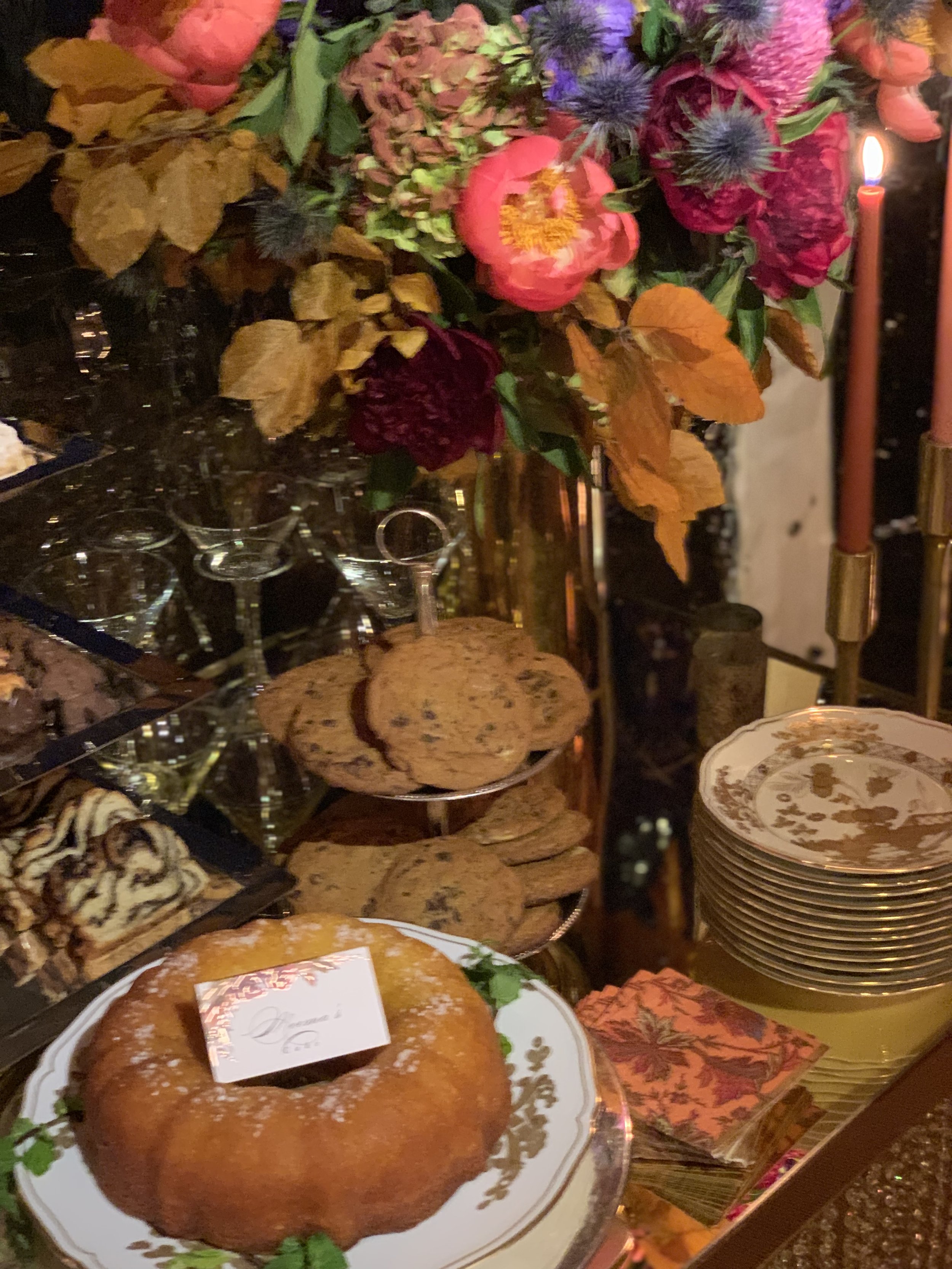 Festive buffet table with colorful flowers, lit candles, cookies, pastries, stacked plates, and wine glasses.
