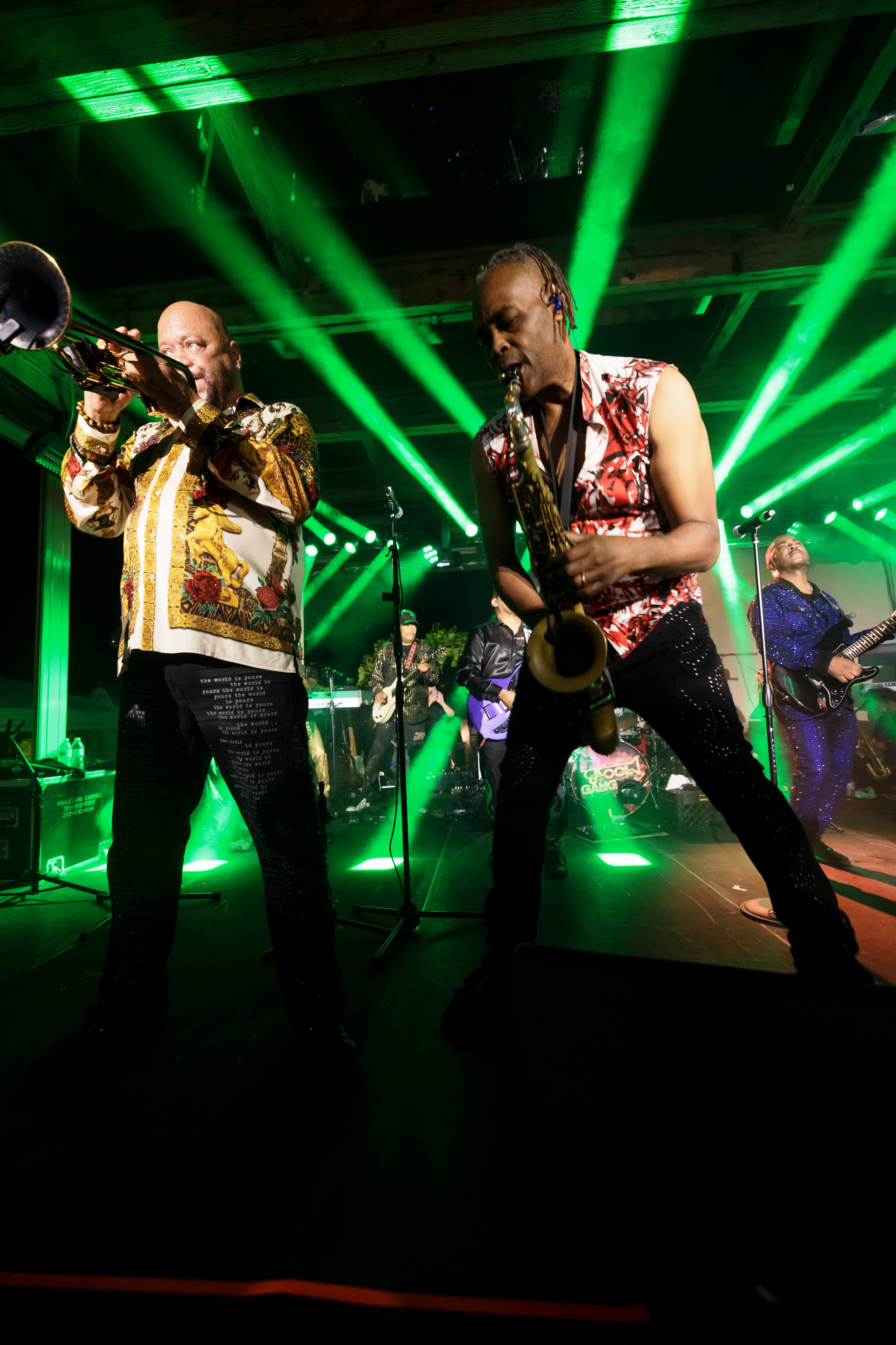 Live band performing onstage under green lights, featuring musicians playing trumpet, saxophone, guitar, and drums.