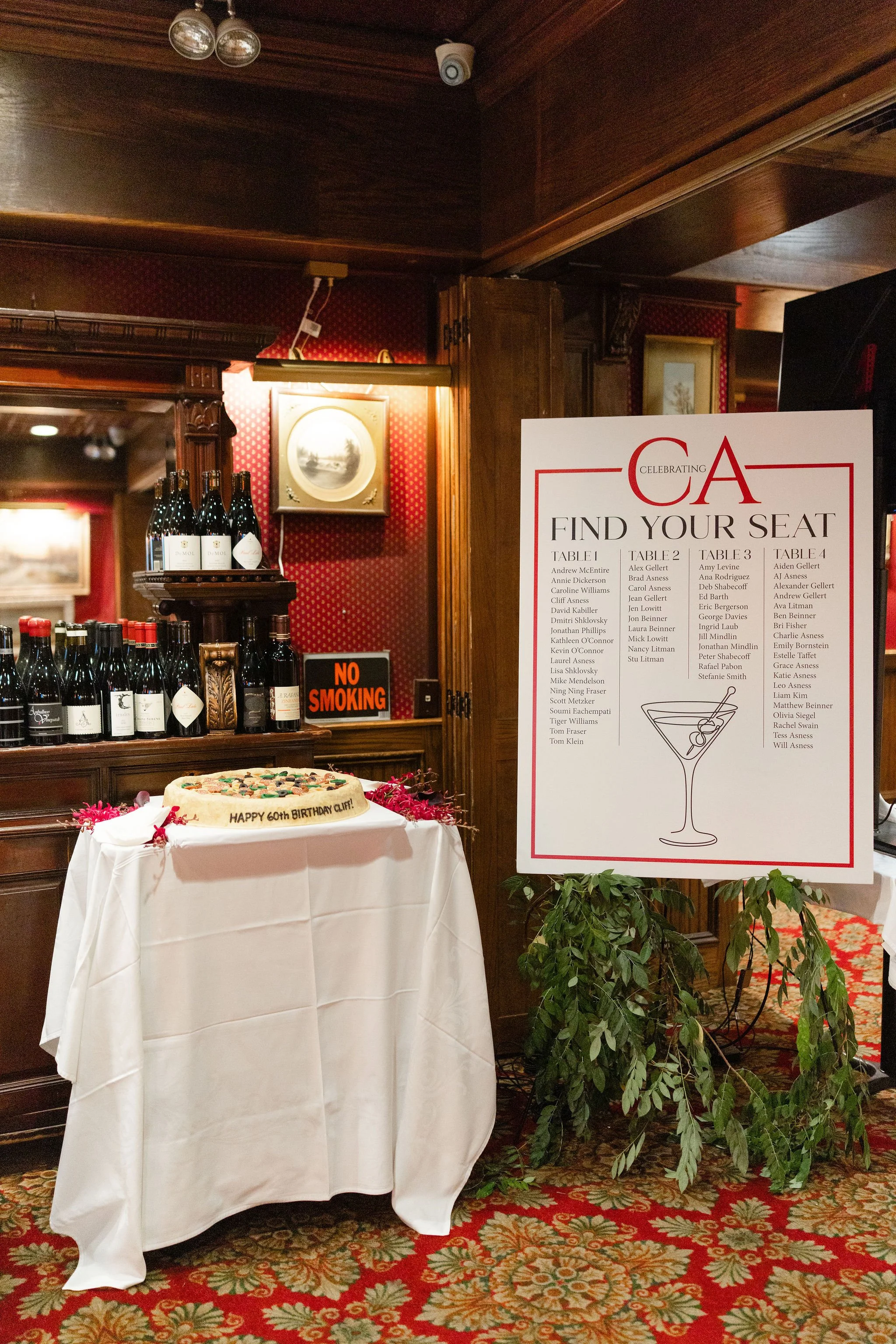 Birthday cake table with wine bottles and a seating chart inside an ornate wood-paneled restaurant
