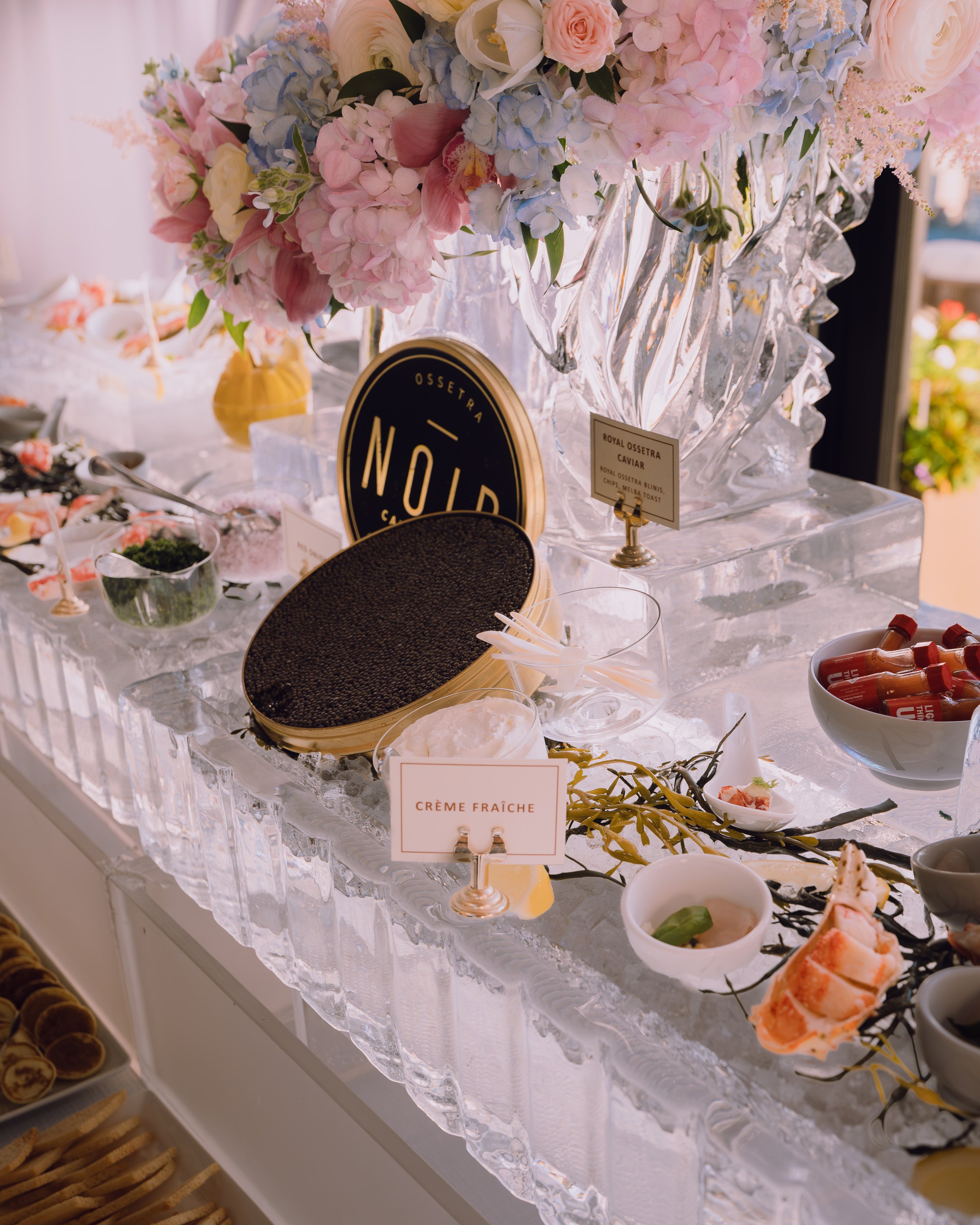 Elegant caviar station with black caviar, crème fraîche, blinis, seafood appetizers, and pastel floral arrangements on ice.
