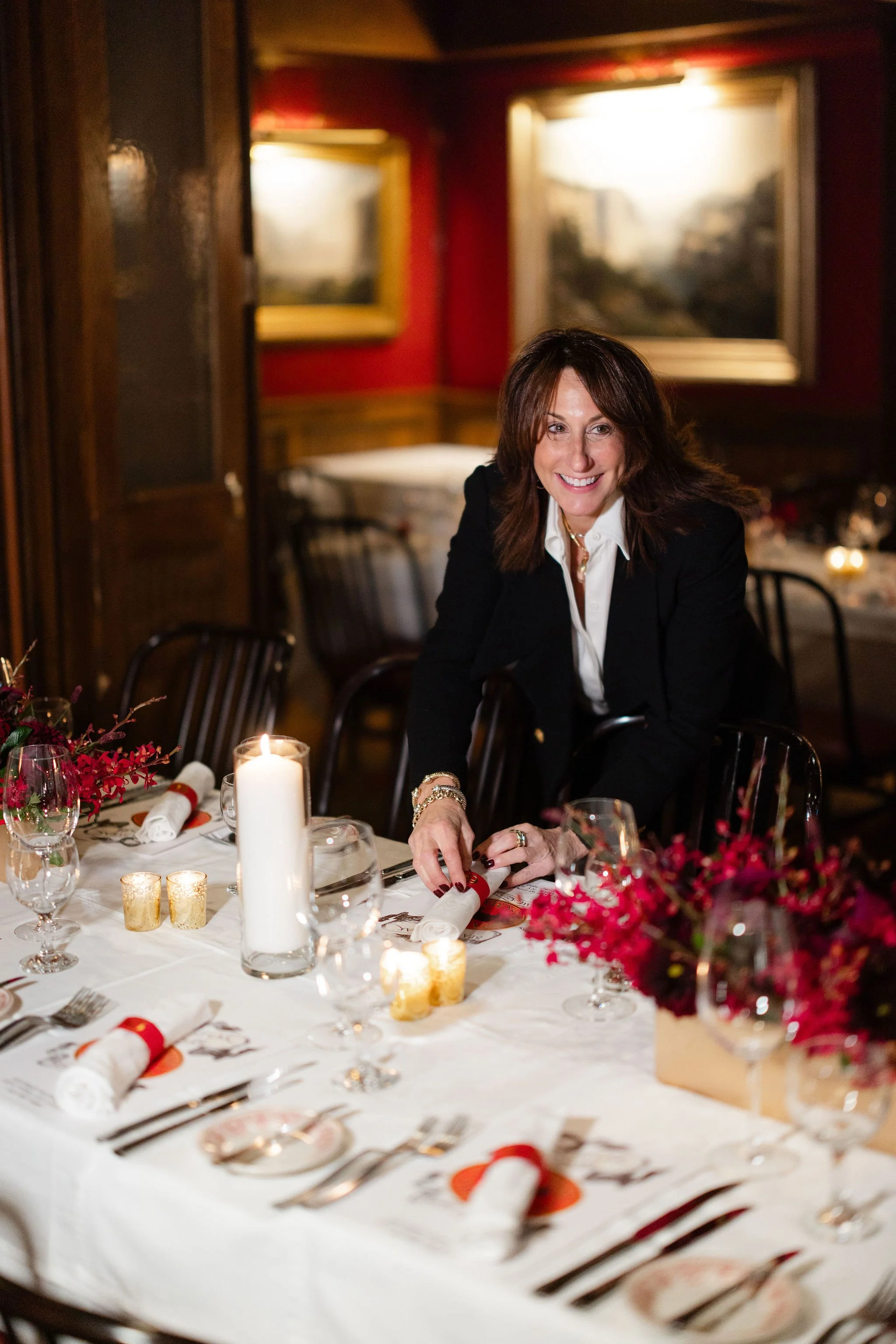 Smiling woman in a black blazer seated at a candlelit restaurant table with floral centerpieces.