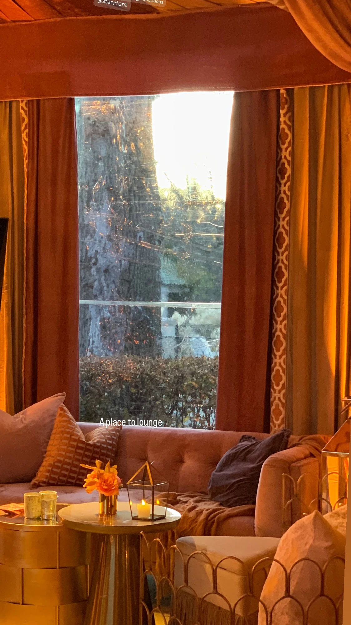 Cozy lounge with a tufted sofa, patterned pillows, candlelit coffee table, and floor-to-ceiling window framed by orange curtains.
