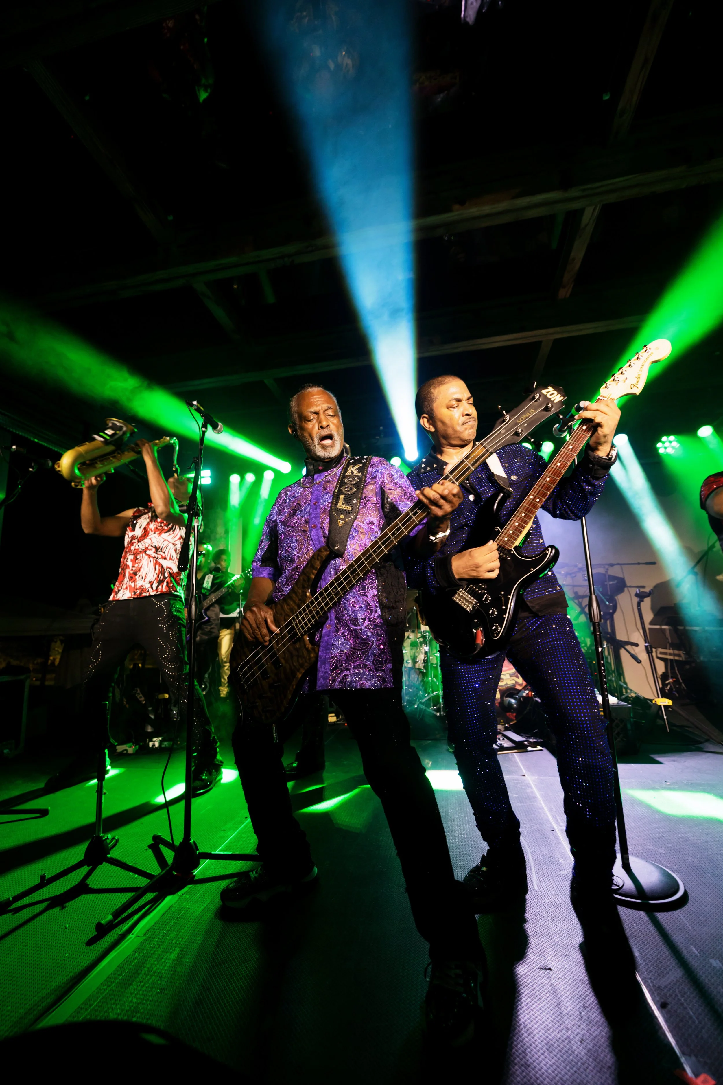 Live band performing onstage with a bassist, guitarist, and saxophonist under green and blue concert lights