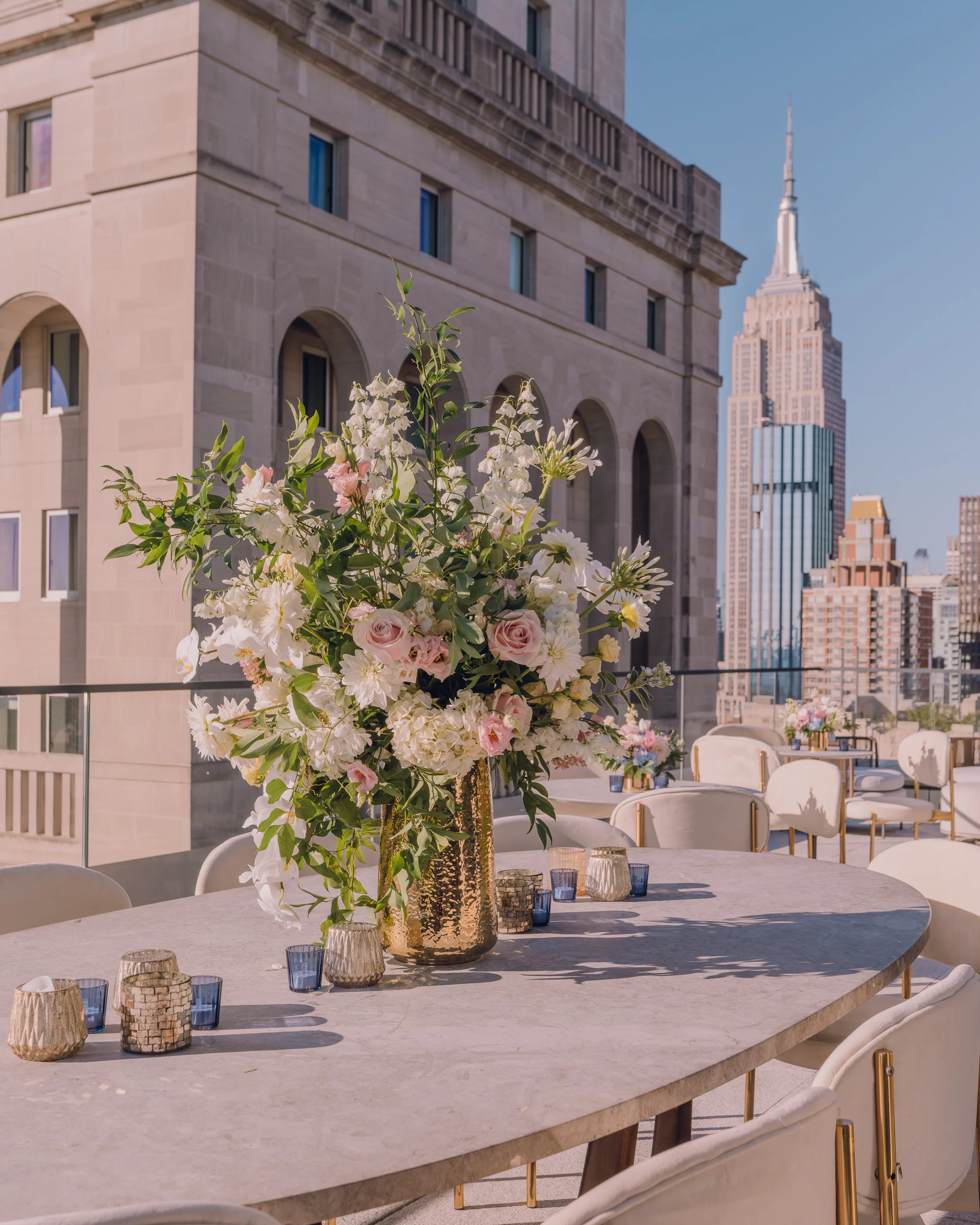 Rooftop dining terrace with a large floral centerpiece, elegant table settings, and a city skyline featuring the Empire State Building.