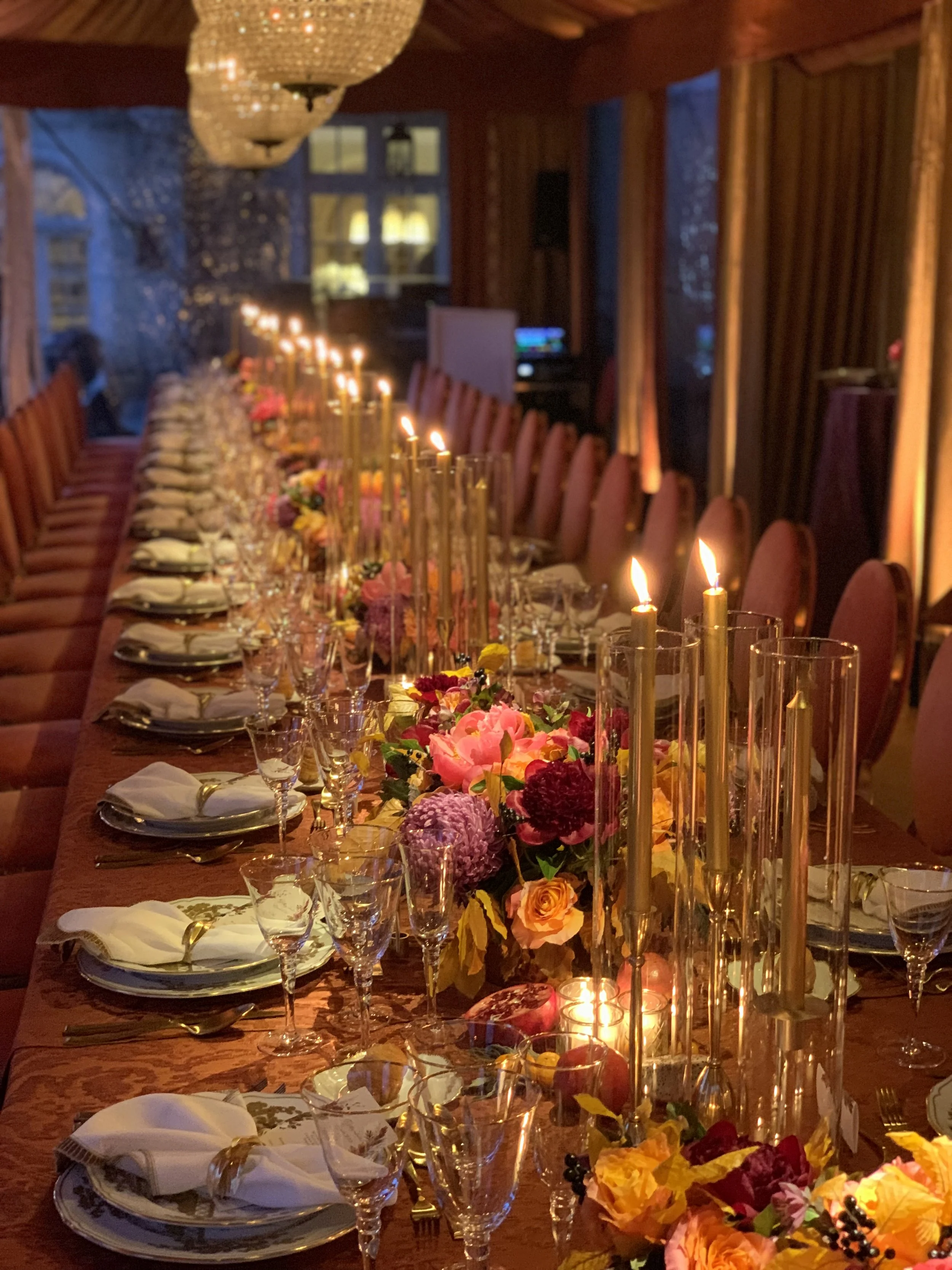 Elegant dinner table set with china plates, wine glasses, floral centerpieces, tall candles, and warm ambient lighting