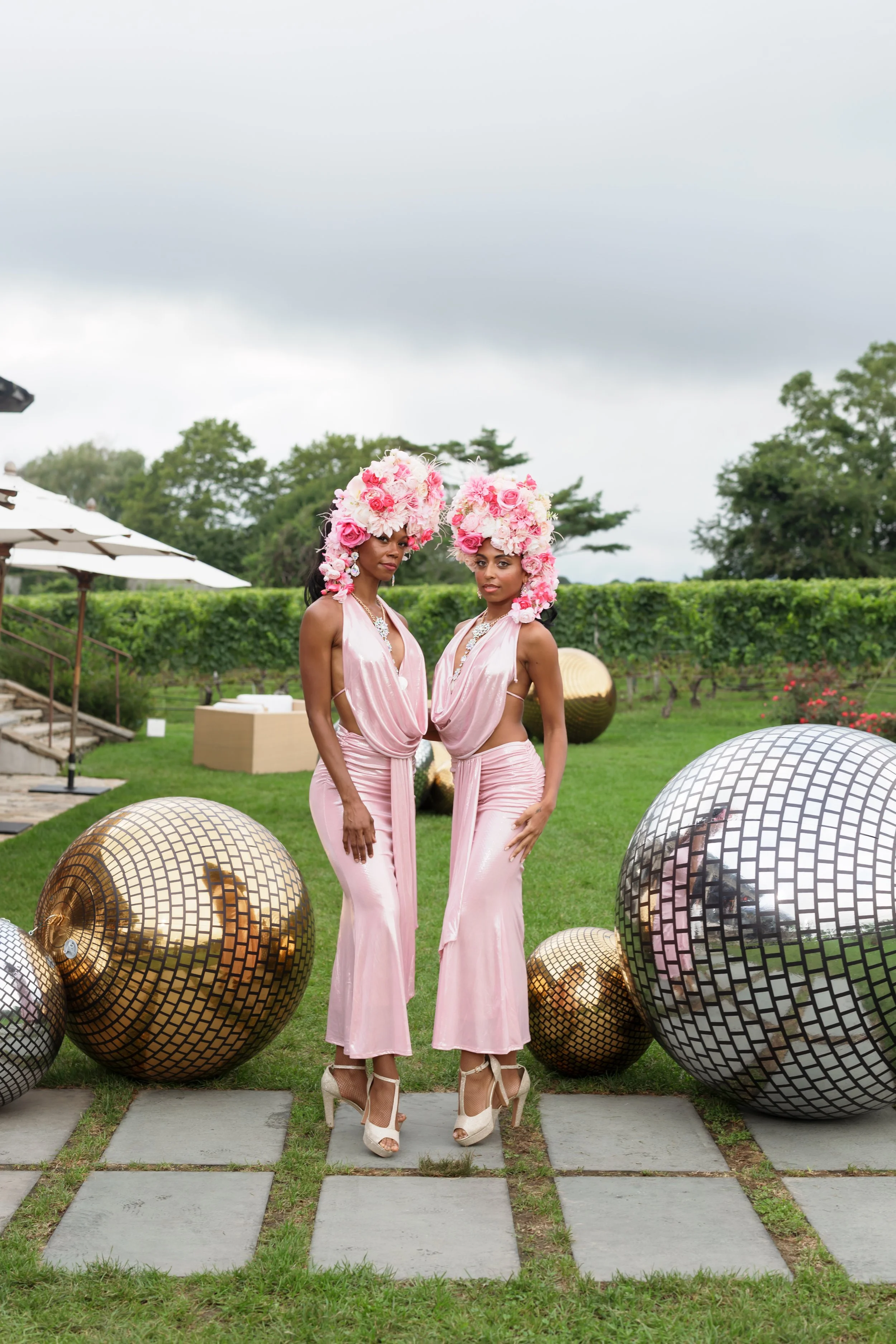 Two women in pink dresses and elaborate floral headpieces pose outdoors among large mirrored disco balls.