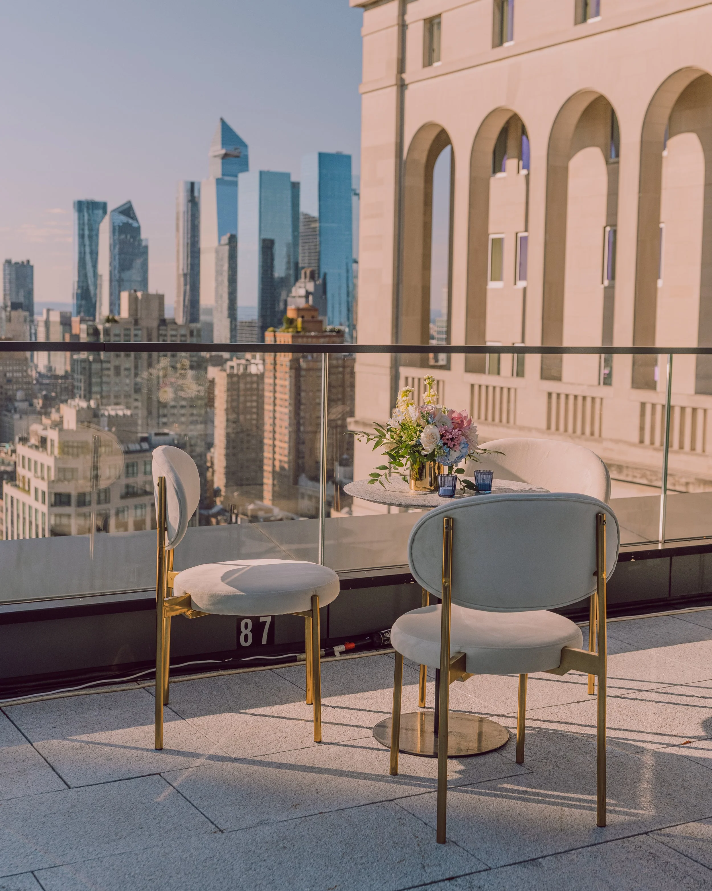 Rooftop terrace dining area with white and gold chairs, a floral centerpiece, and a city skyline view.
