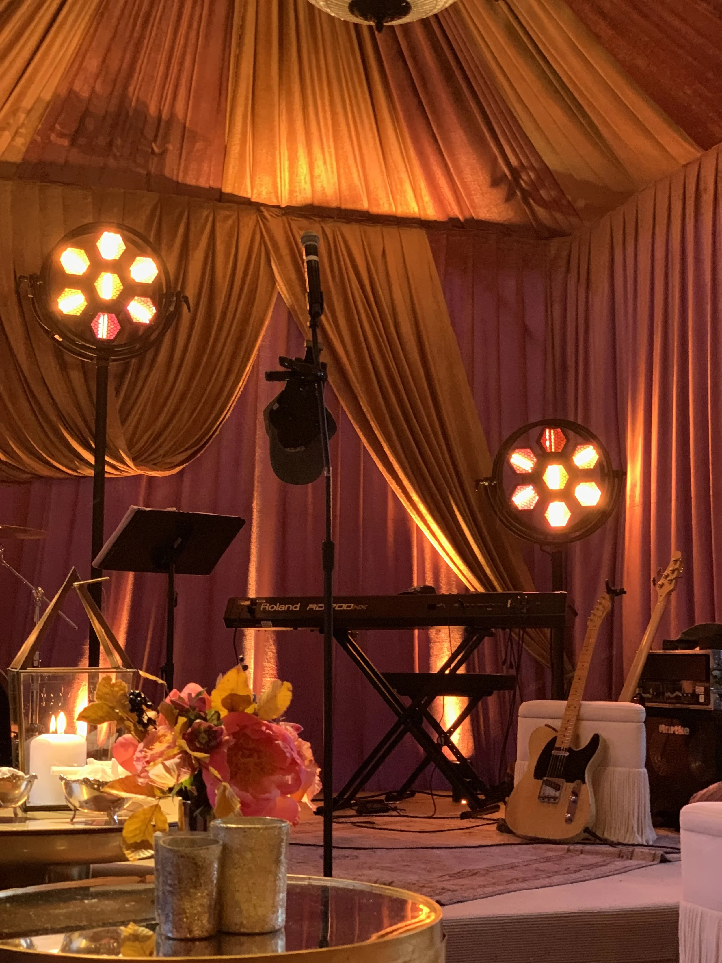 Warmly lit stage with purple and orange drapery, a Roland keyboard, microphone, guitars, theatrical lights, candles, and floral arrangements.