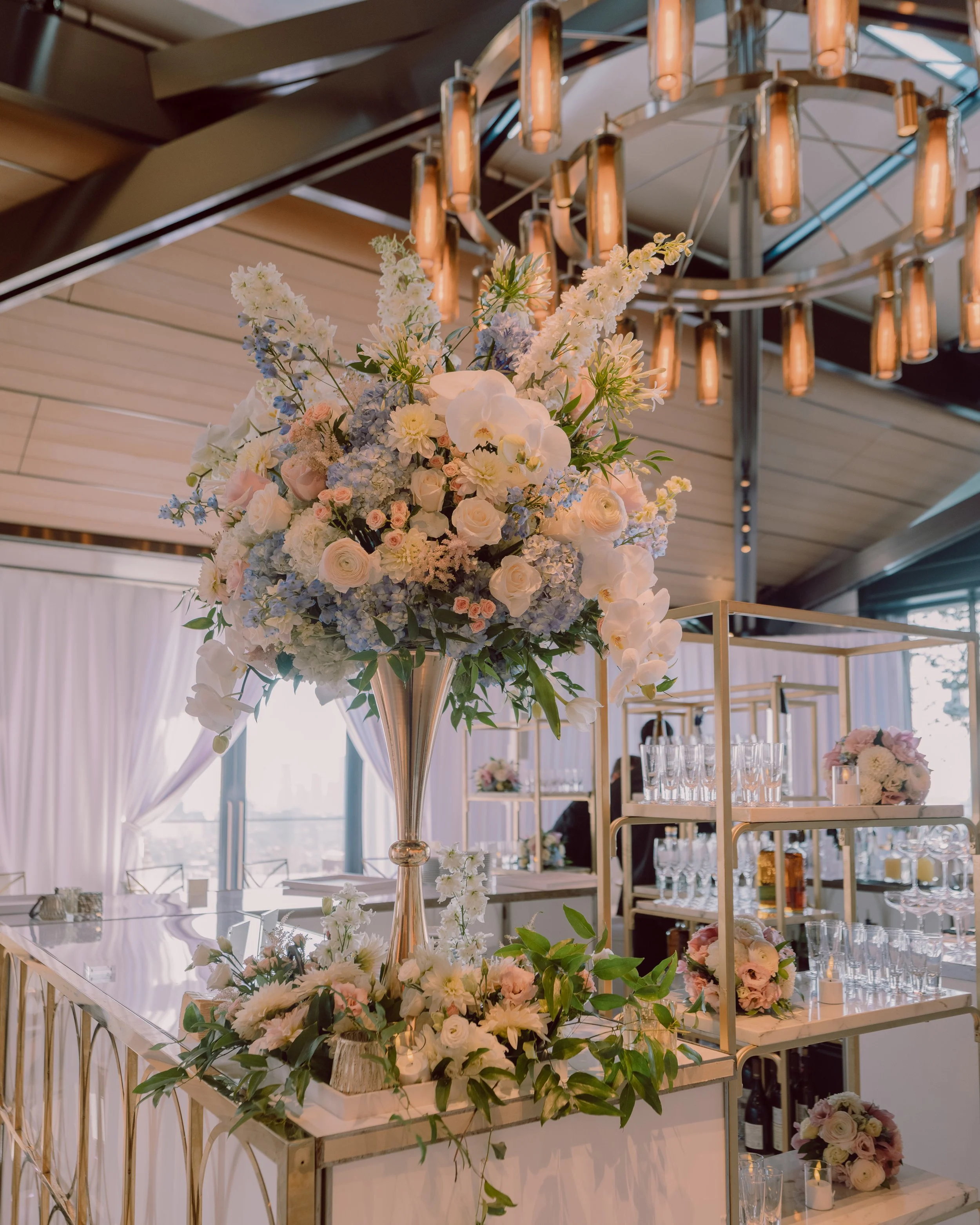 Elegant wedding reception with a large pastel floral arrangement, gold hanging lights, and a champagne bar with glassware.