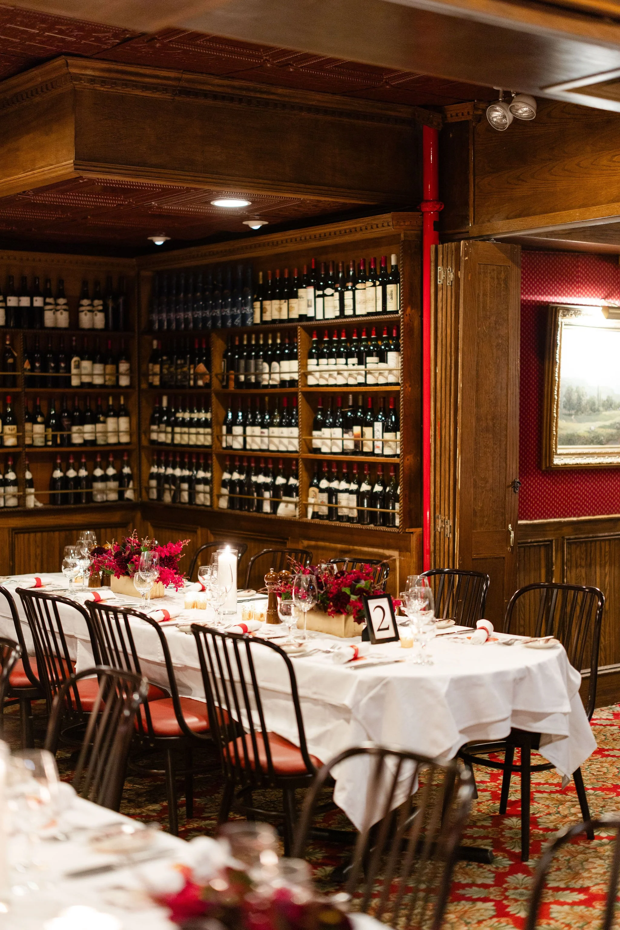 Elegant restaurant dining room with wine-filled wooden shelves, white tablecloths, floral centerpieces, and set tables.