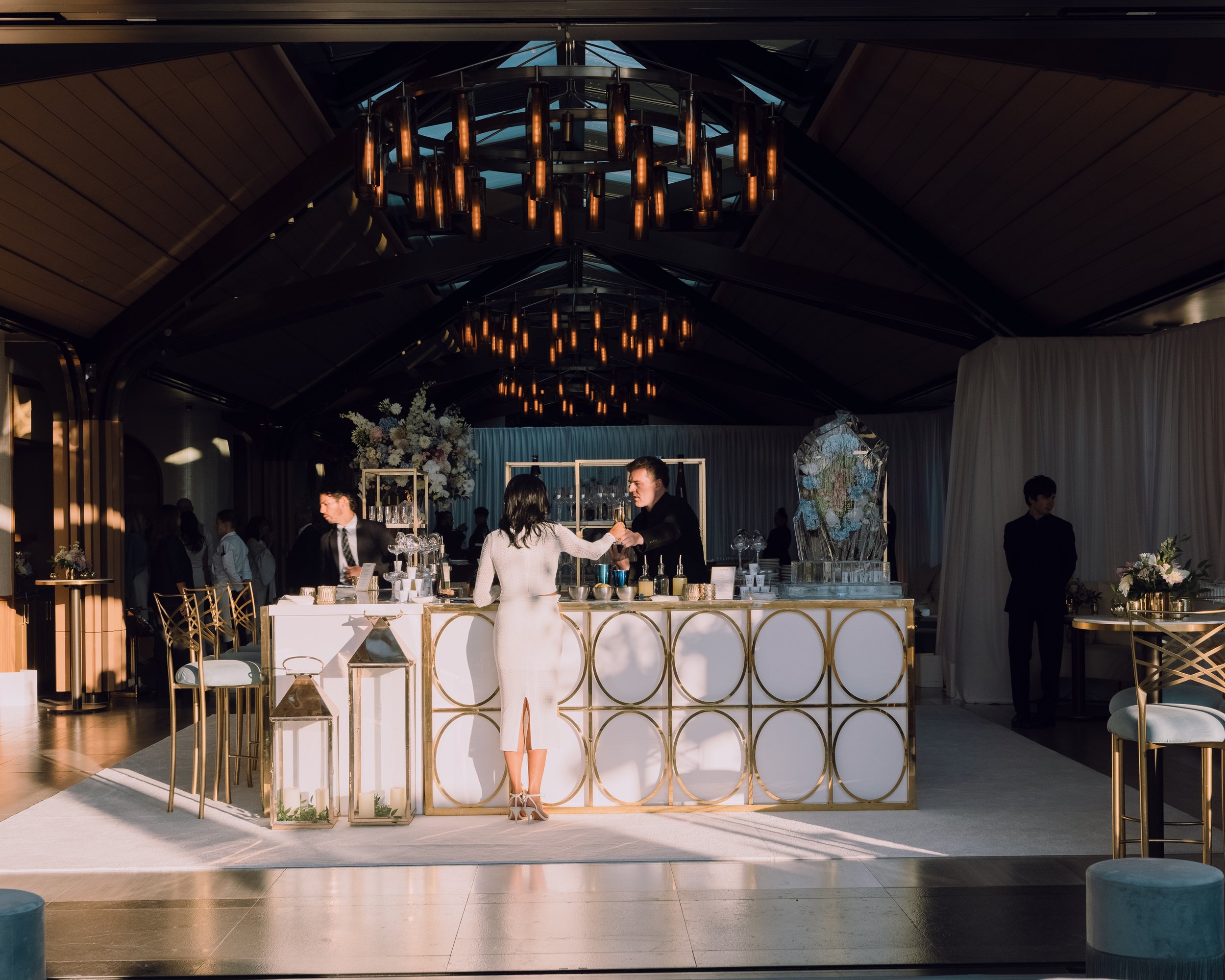 Elegant event bar with a white and gold geometric counter, bartenders, guests, floral arrangements, and a large ice sculpture beneath dramatic pendant lights.